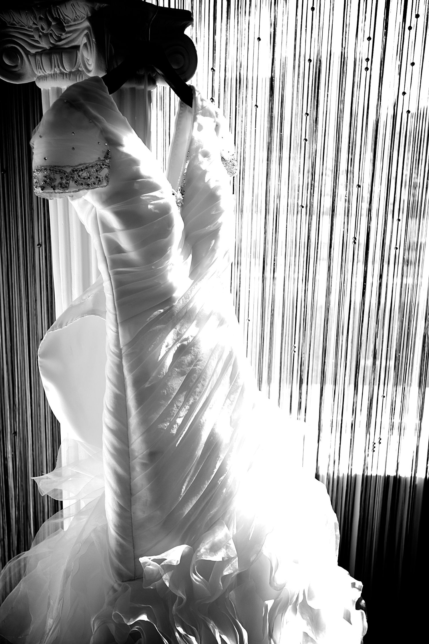 Black and white photo of an elegant wedding dress displayed near a curtained window, showcasing intricate details and flowing fabric.