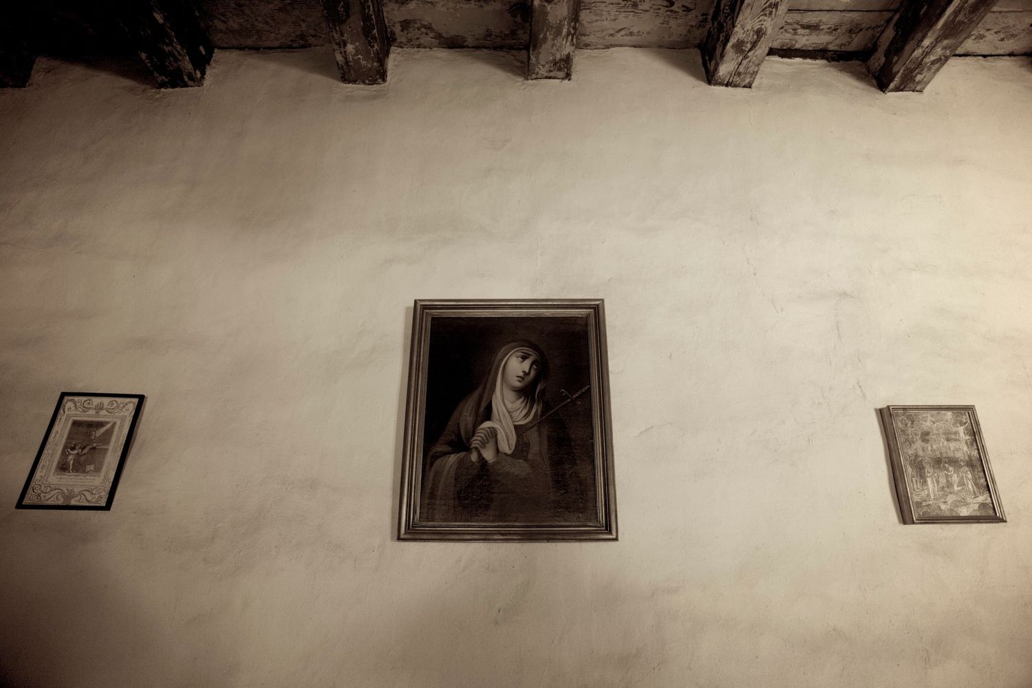 Sepia-toned photo of antique religious paintings on a weathered white wall with wooden ceiling beams.