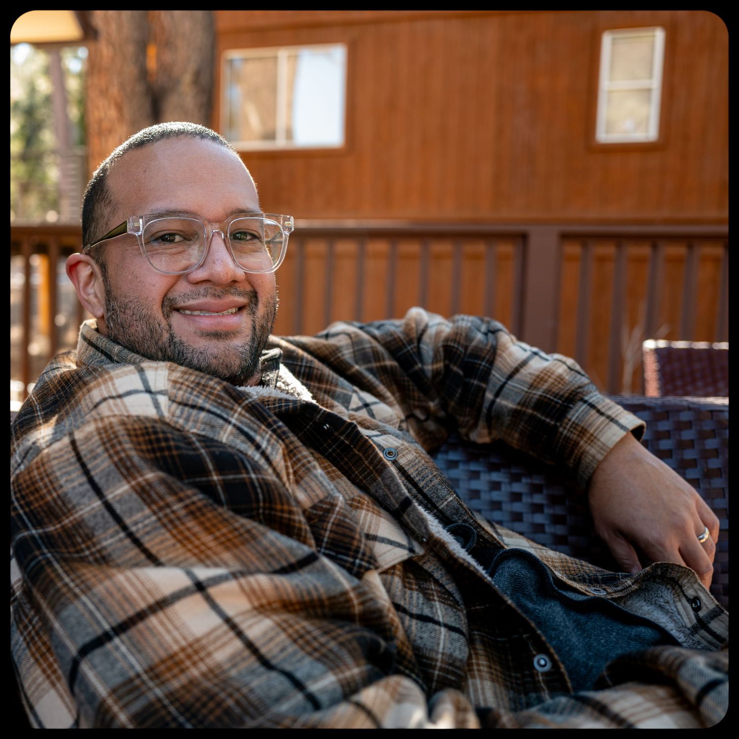 A smiling man with glasses wearing a plaid flannel shirt relaxes in a chair on a wooden deck cabin porch.