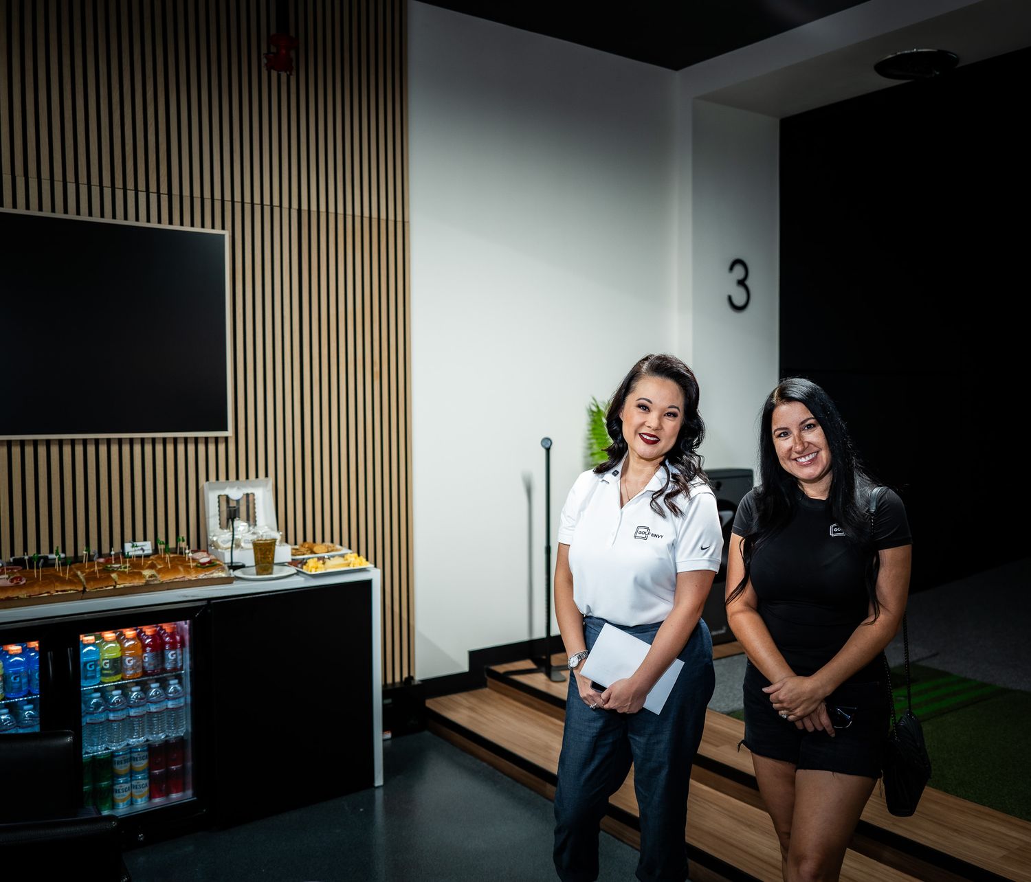 Two smiling women in matching white and black outfits pose in a modern room with a wood slat wall, TV, and a refreshment bar with drinks.