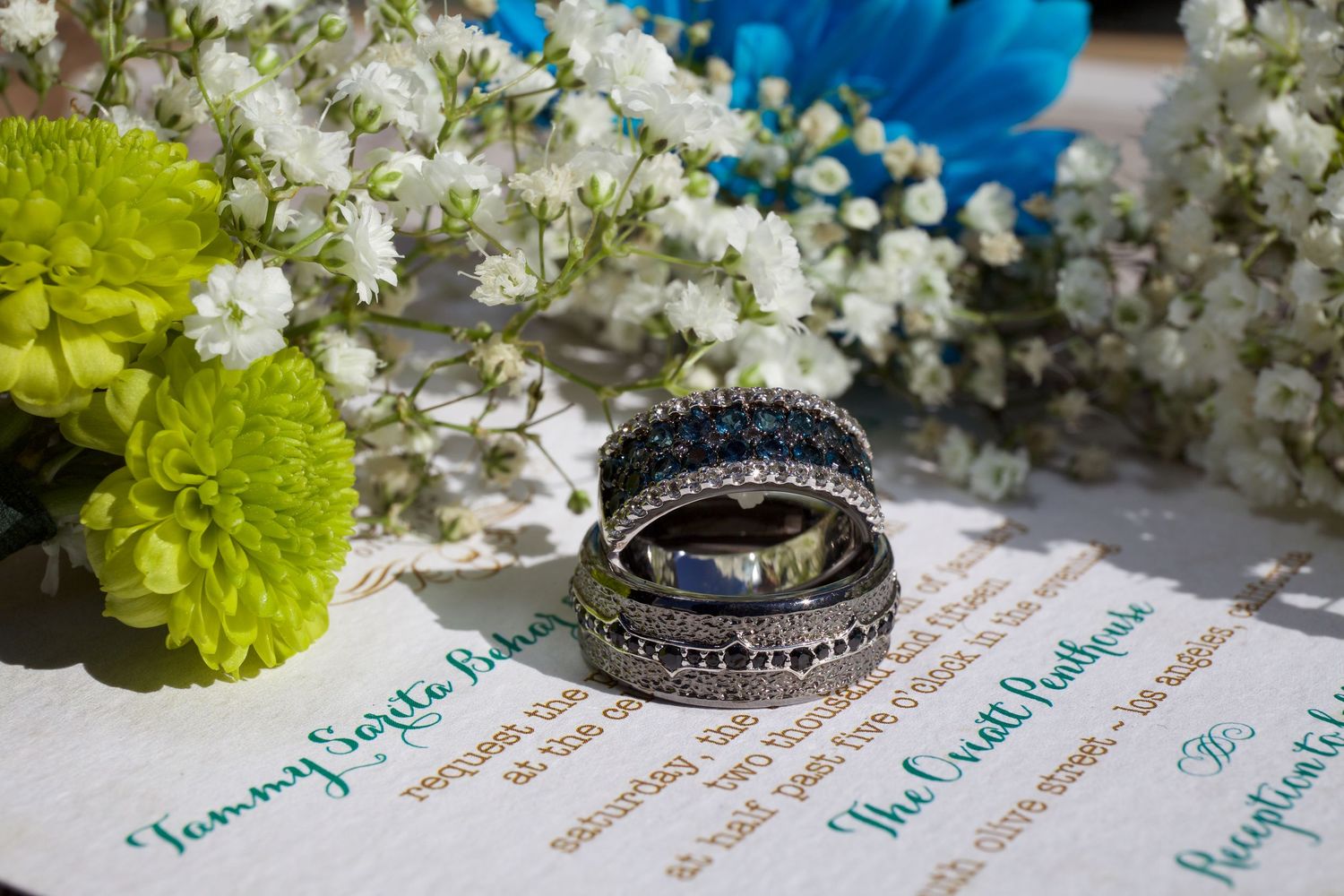 Close-up of stacked wedding bands with sapphire accents resting on wedding invitation with floral decor.
