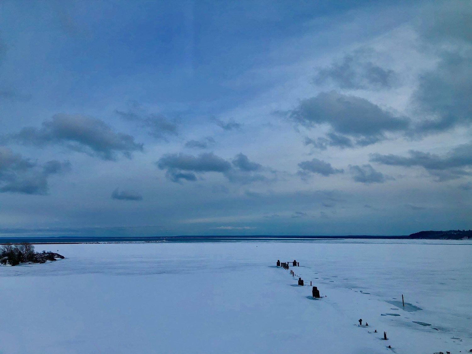 Frozen lake under cloudy blue winter sky with snow-covered ice stretching to distant treeline.