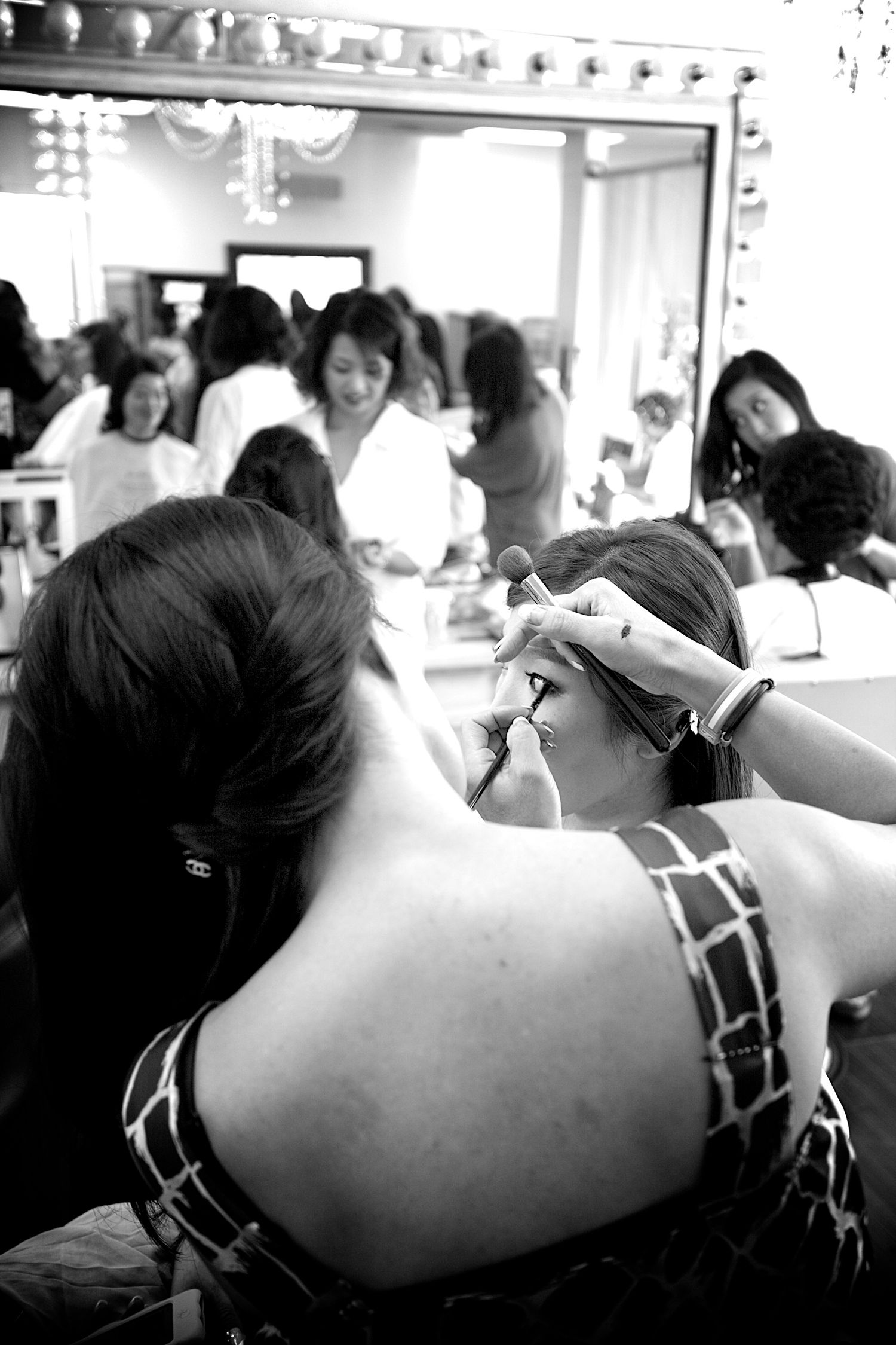 Black and white photo of women in a busy dressing room, one styling another's hair before an event.