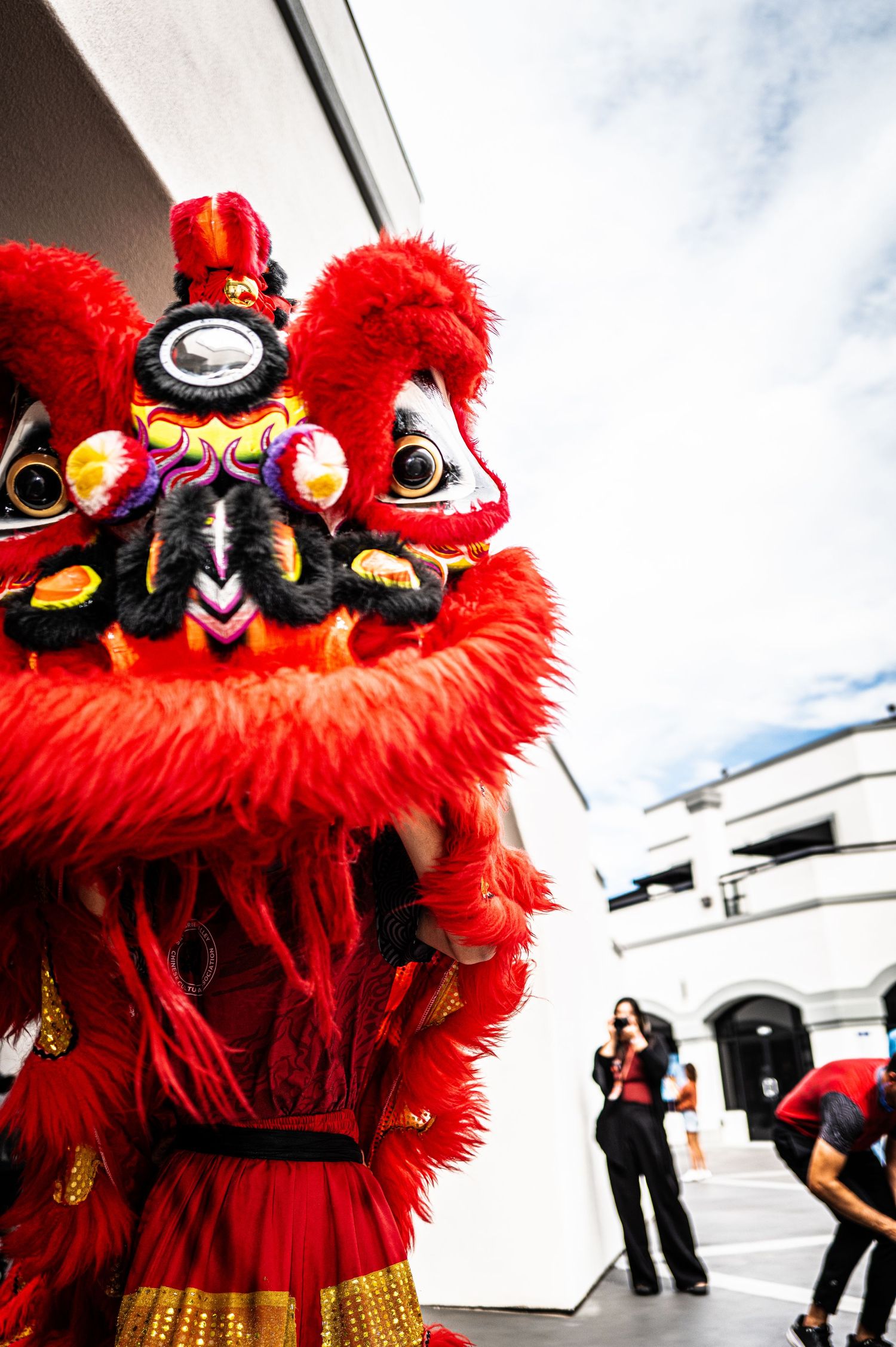 Red and gold lion dance costume with fuzzy fur and ornate eyes performing outdoors as spectators stand near a white building.