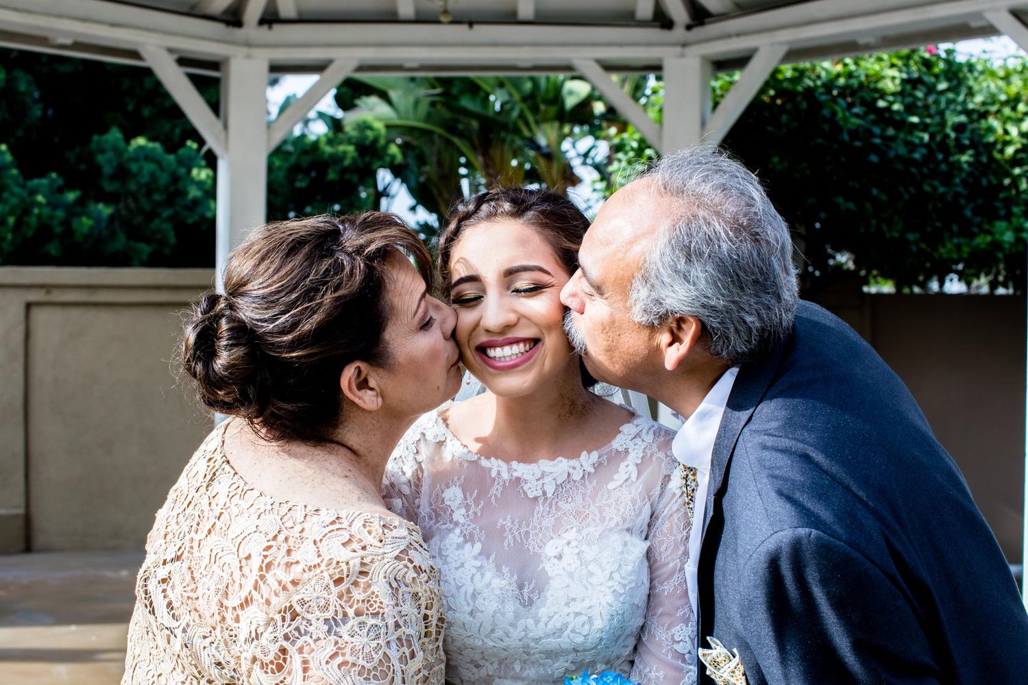 Bride in white lace gown smiles joyfully as parents kiss her cheeks during wedding celebration outdoors.