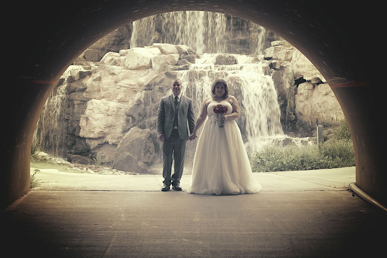 Bride and groom holding hands under stone arch tunnel with waterfall backdrop on their wedding day.