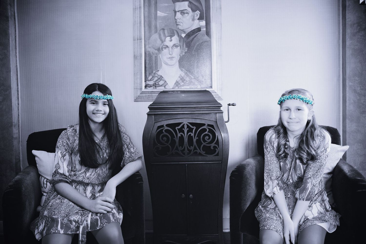 Two young girls wearing floral headbands seated in chairs in a black and white artistic photograph.