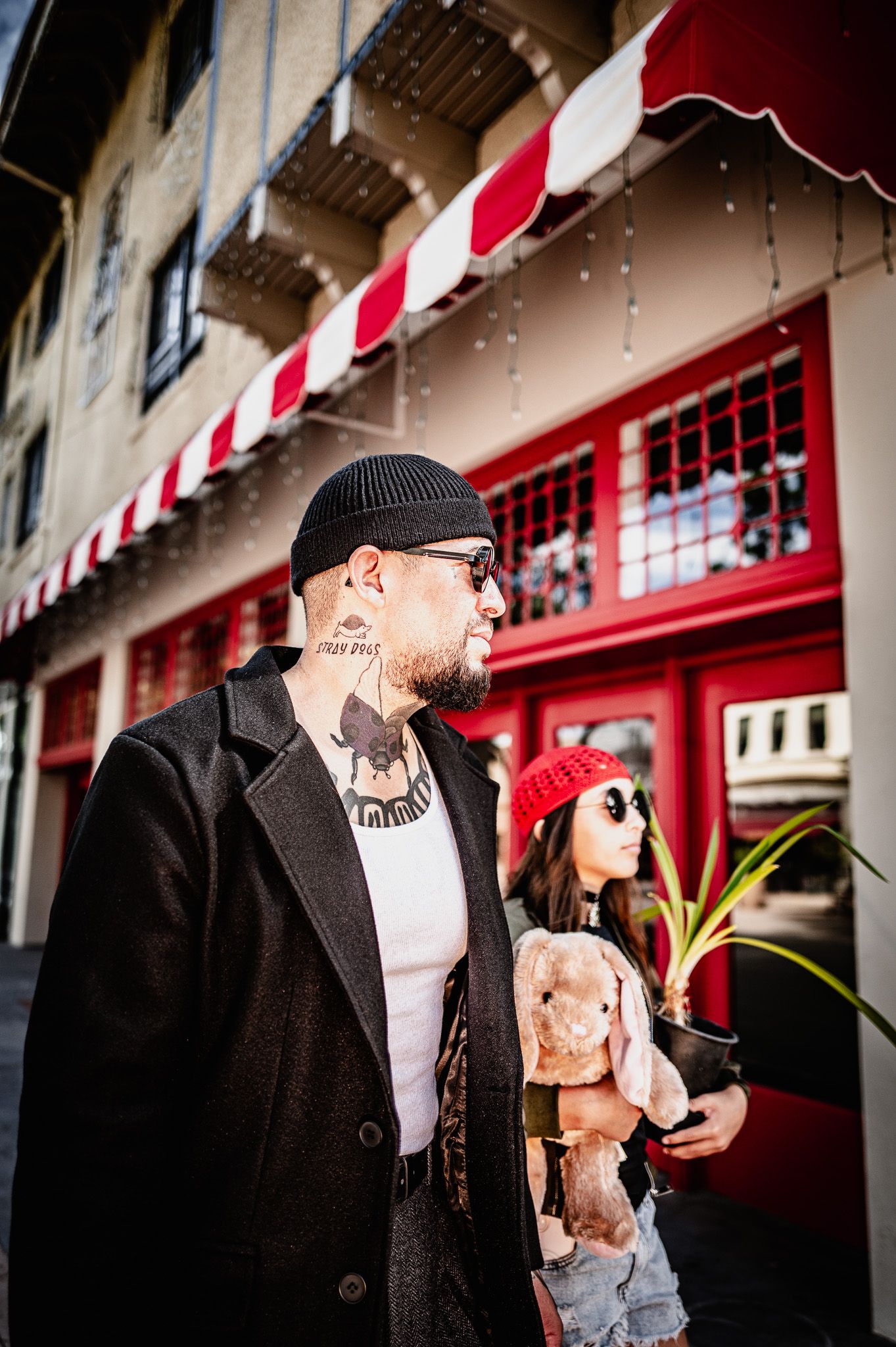 Bearded man with neck tattoos, beanie and sunglasses walks beside a girl in a red hat holding a plush bunny and plant by a red storefront.