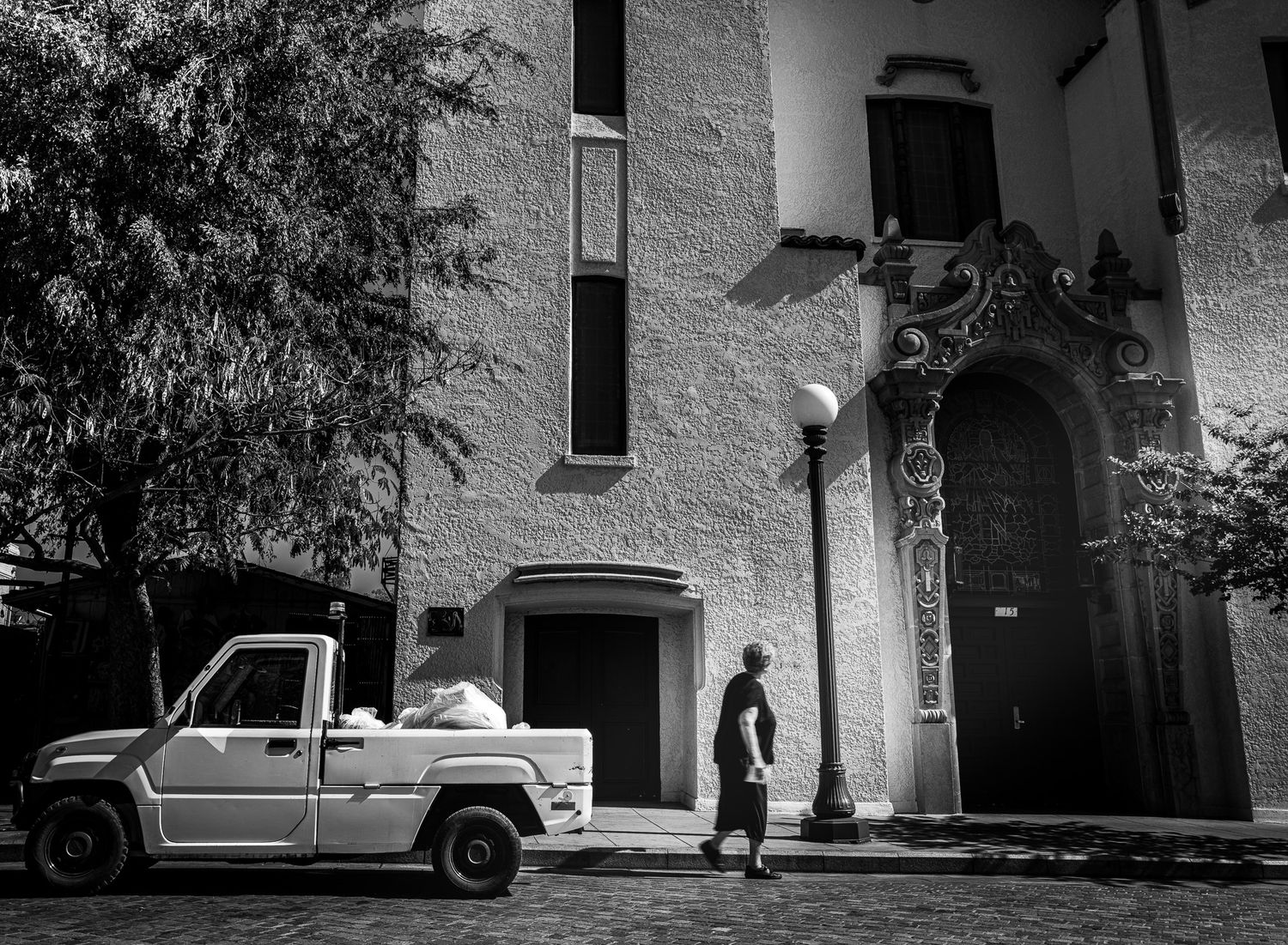 Black and white street photo of a man walking past a pickup truck near an ornate building entrance with a street lamp.