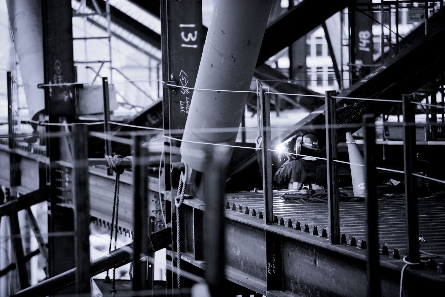 Black and white industrial scene showing steel beams and a welder working in a large structure.