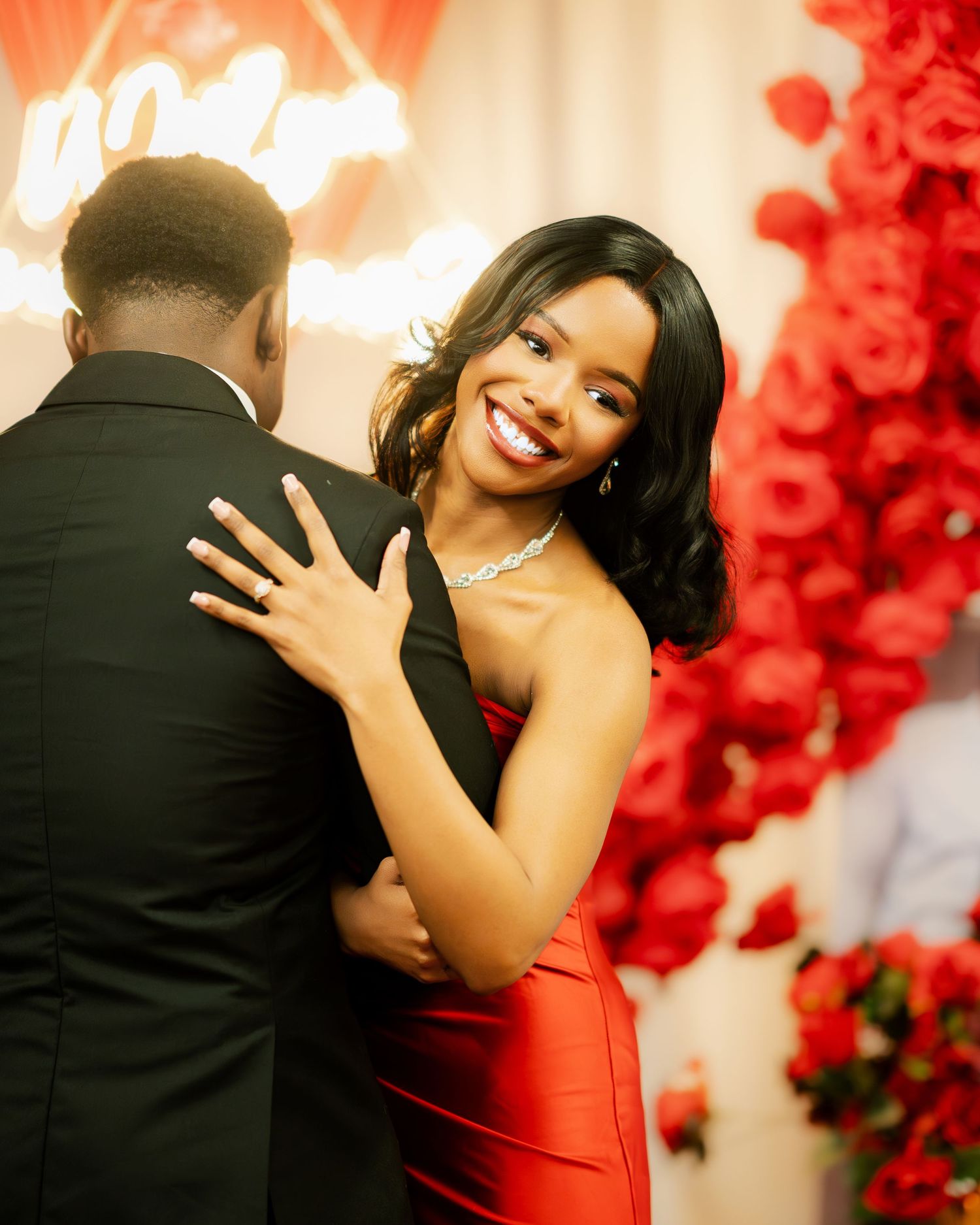A woman in a red strapless gown smiling radiantly while embracing a man in a black suit at a romantic proposal setup.