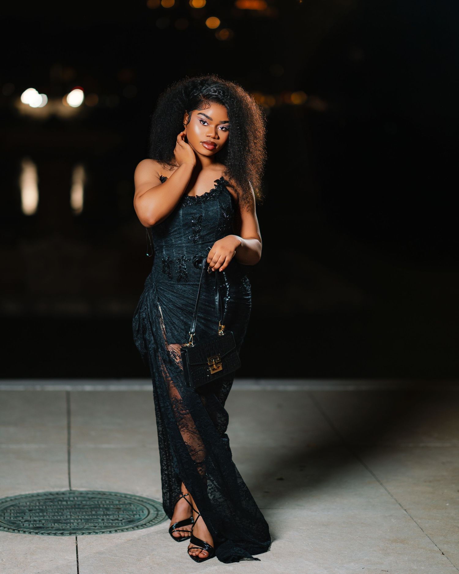 Stylish young woman in black lace maxi dress posing outdoors at night in strappy heels.