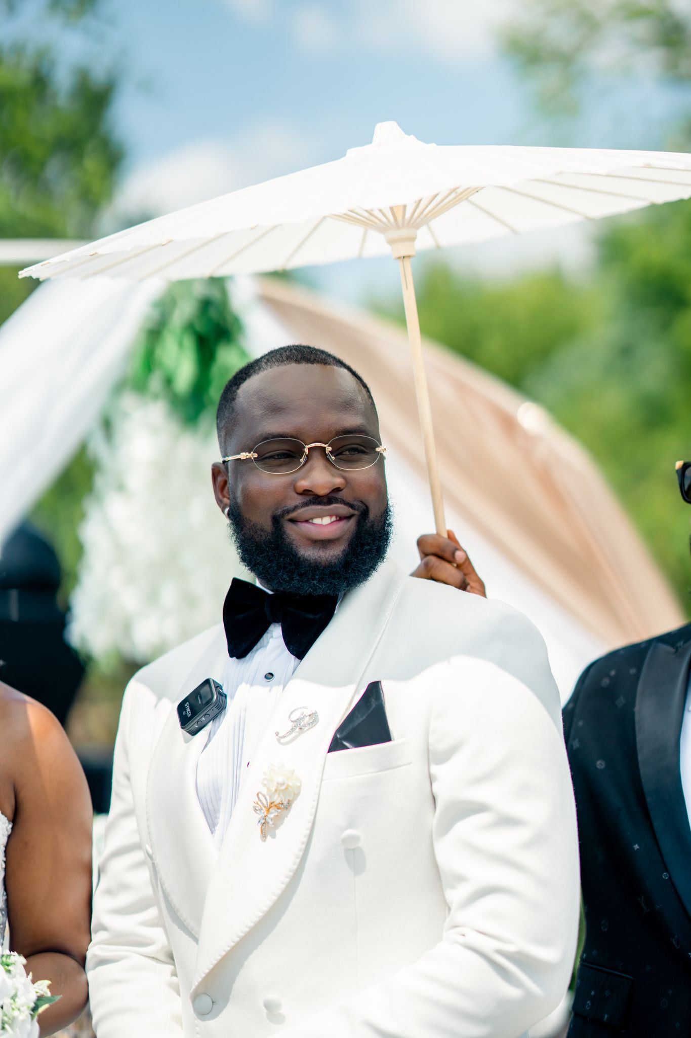 A handsome groom in a white tuxedo smiles while holding a white parasol during an outdoor wedding ceremony.
