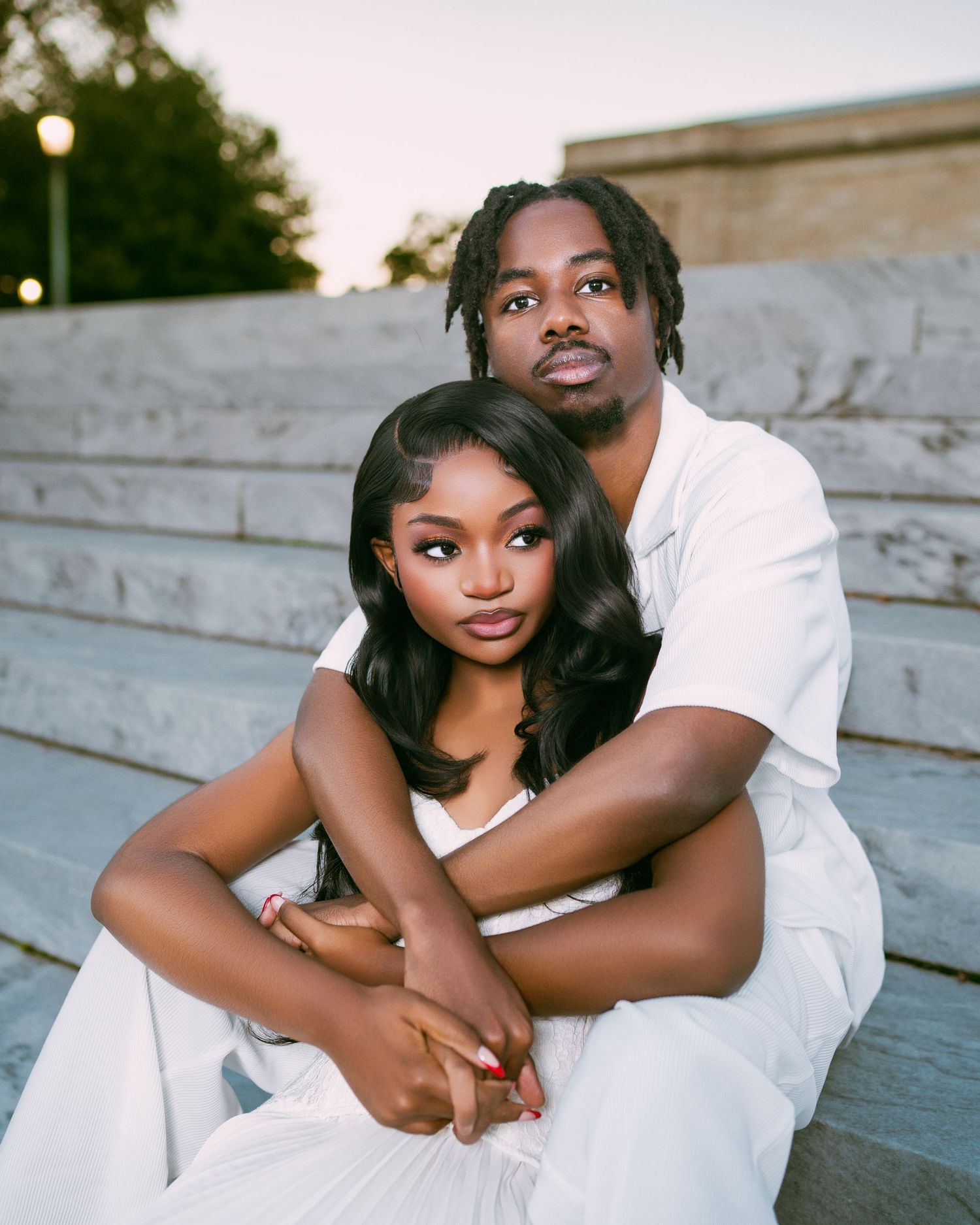 A young Black couple sits on stone steps wearing matching white outfits in a romantic outdoor portrait.