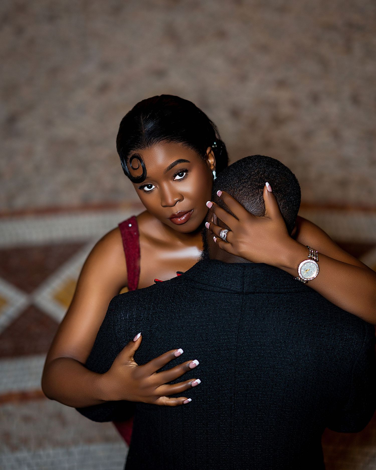 Woman in burgundy dress embracing man in black suit, showcasing diamond engagement ring.