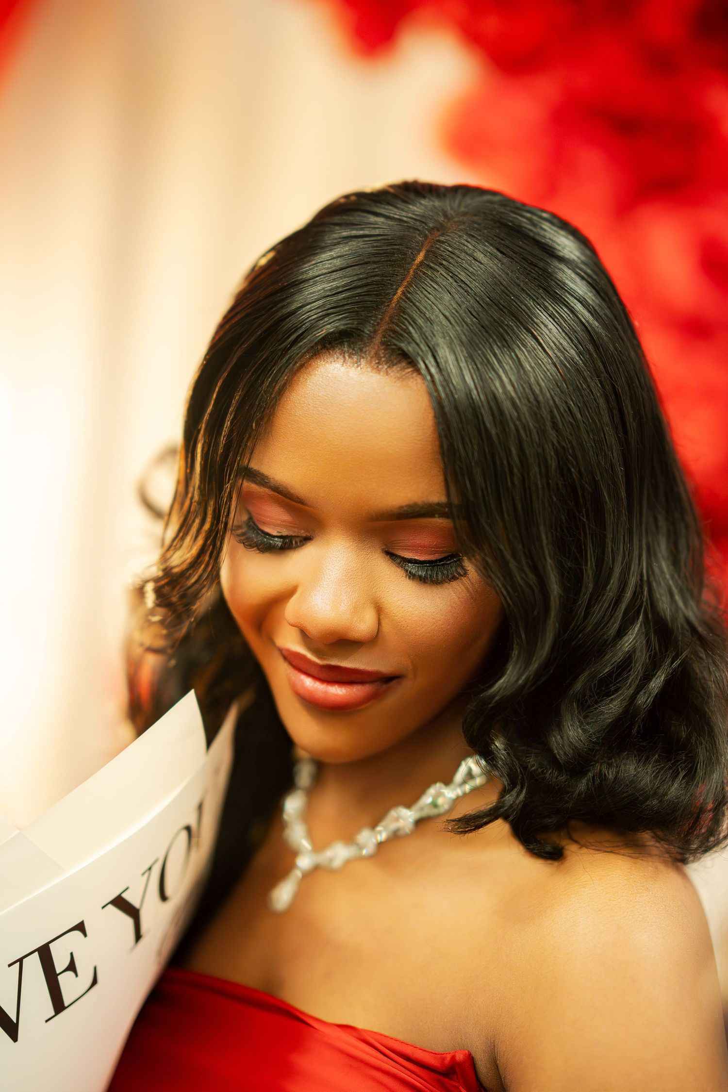 An elegant woman in a red strapless gown with a diamond necklace reads an I Love You card at a proposal.