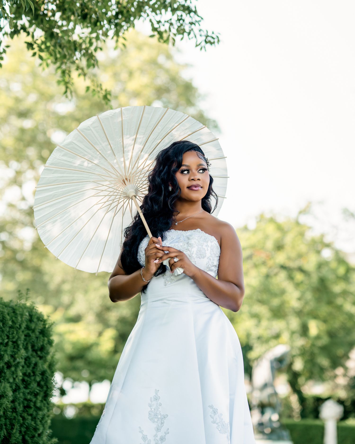 Elegant bride in white strapless wedding gown holds ivory parasol outdoors among green trees.