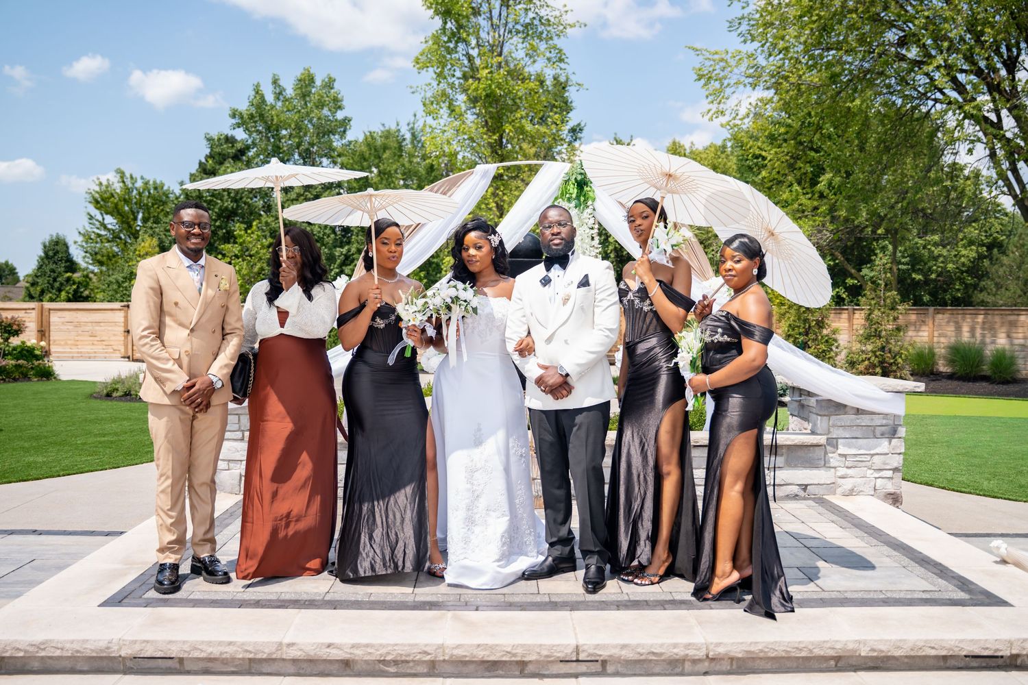 Wedding party posing with white parasols outdoors, bride in white gown and bridesmaids in black dresses.