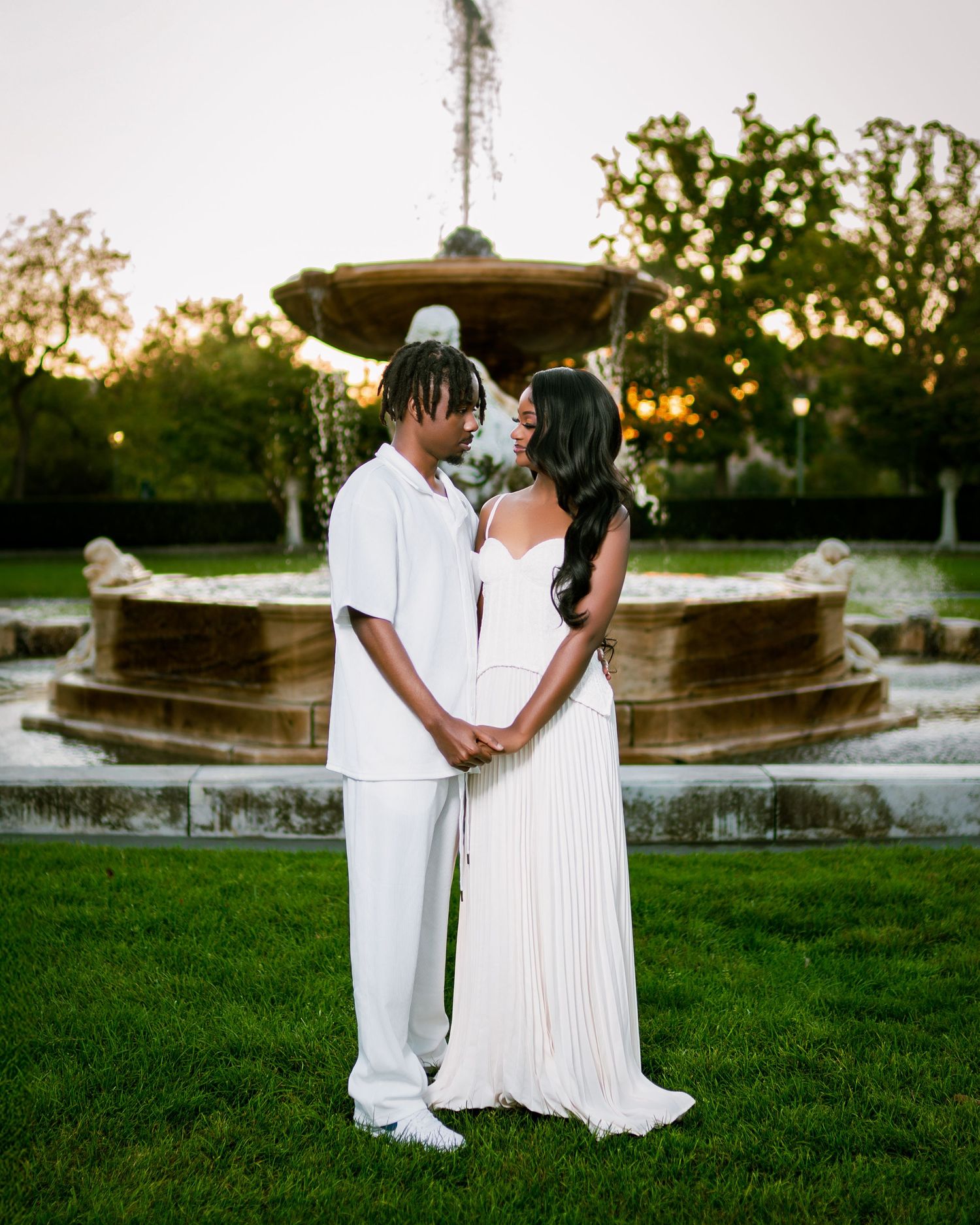 Couple in white outfits holding hands and gazing at each other before a stone garden fountain.