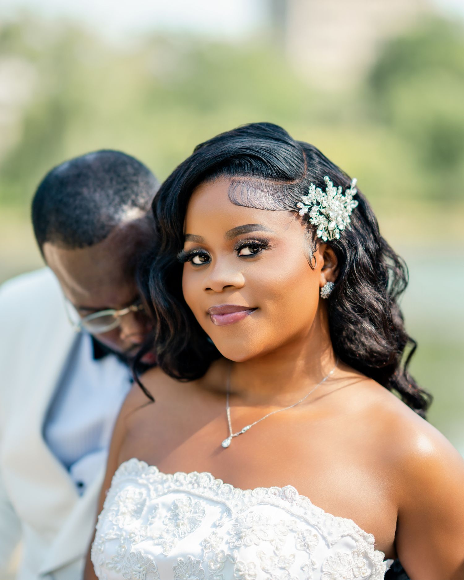 A stunning bride with a jeweled hair accessory gazes at the camera while her groom leans in close behind her.
