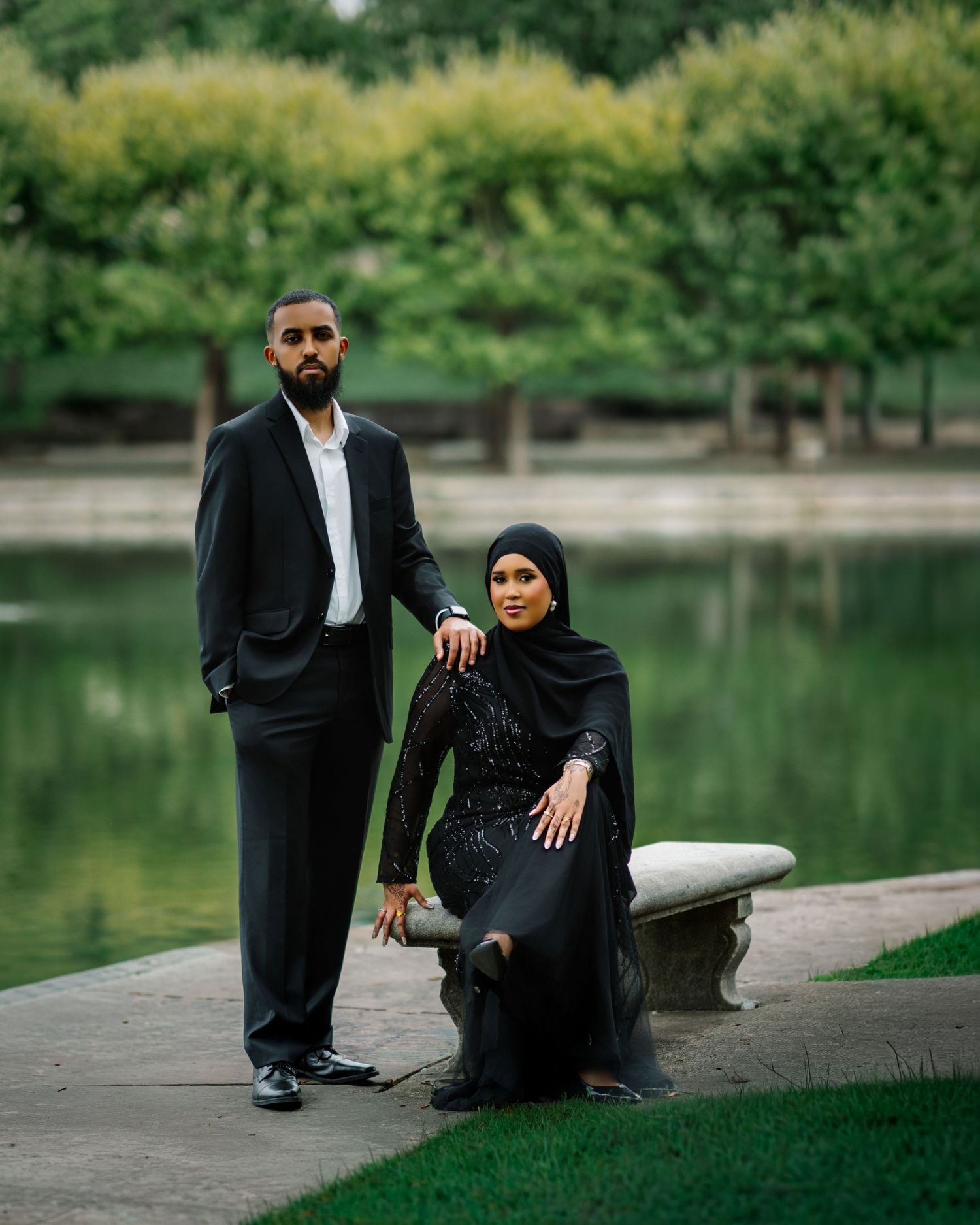 Muslim couple in black formal wear pose by garden pond, woman in hijab seated on stone bench.