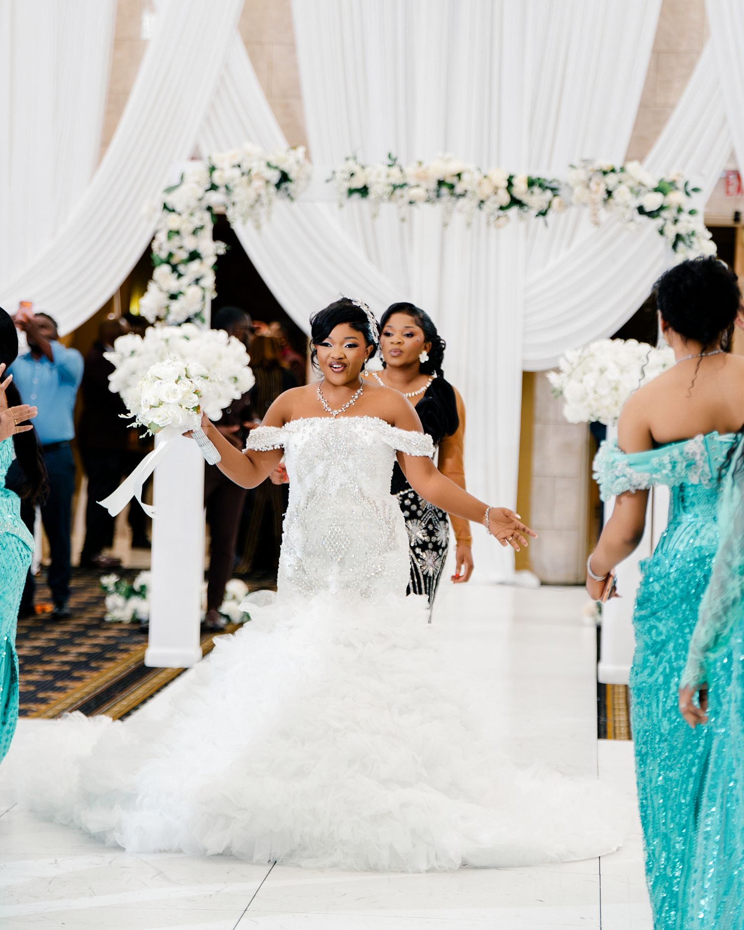 Bride in white mermaid dress walks down floral-decorated aisle with bridesmaids in teal sequin gowns.