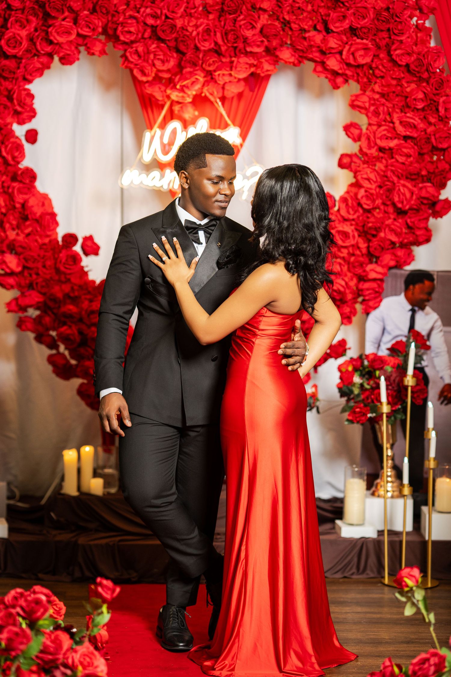 Couple embraces at romantic red rose proposal setup with neon sign and candles in background.