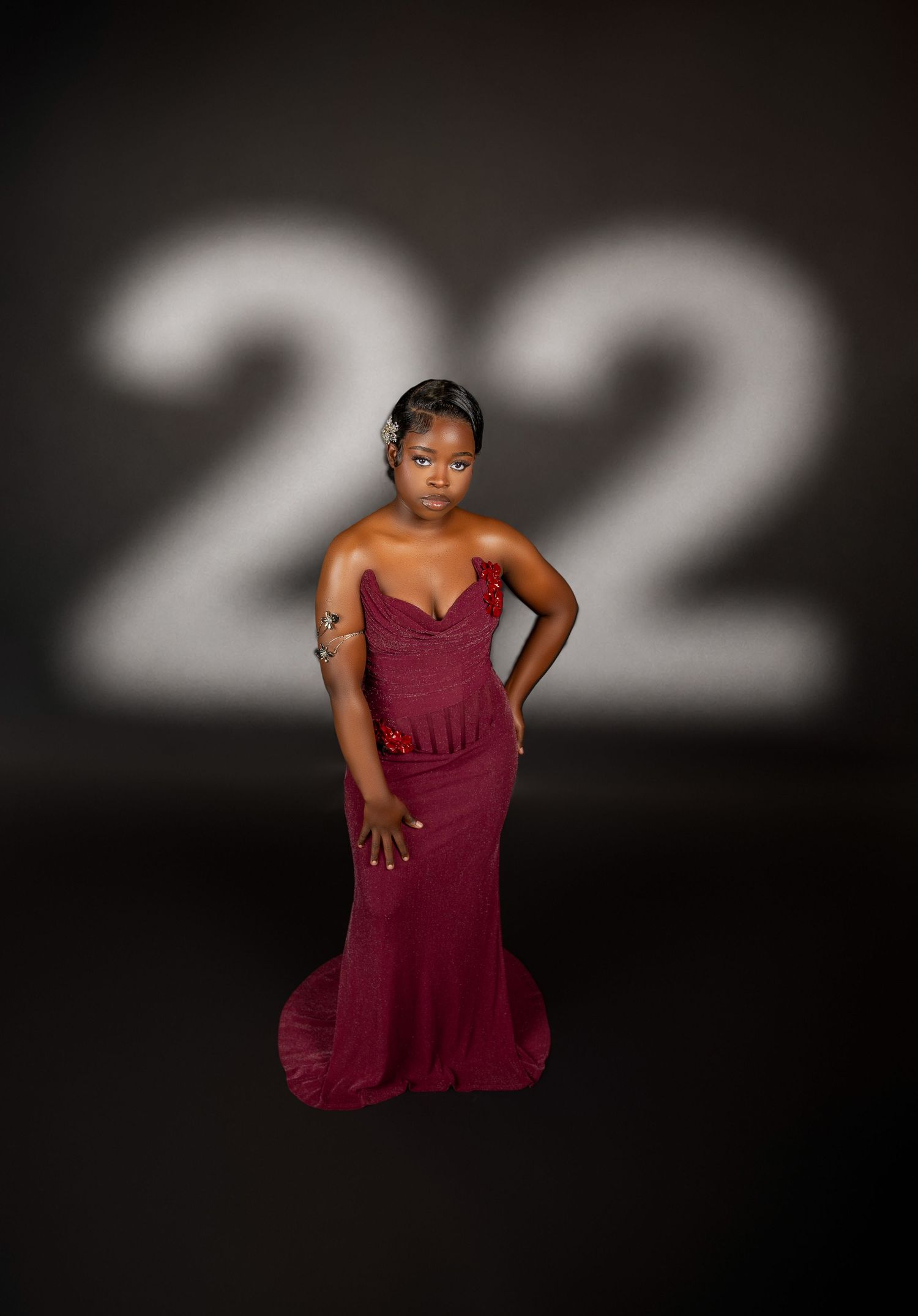 Young woman in a burgundy mermaid gown poses in front of a projected number 22 on dark background.
