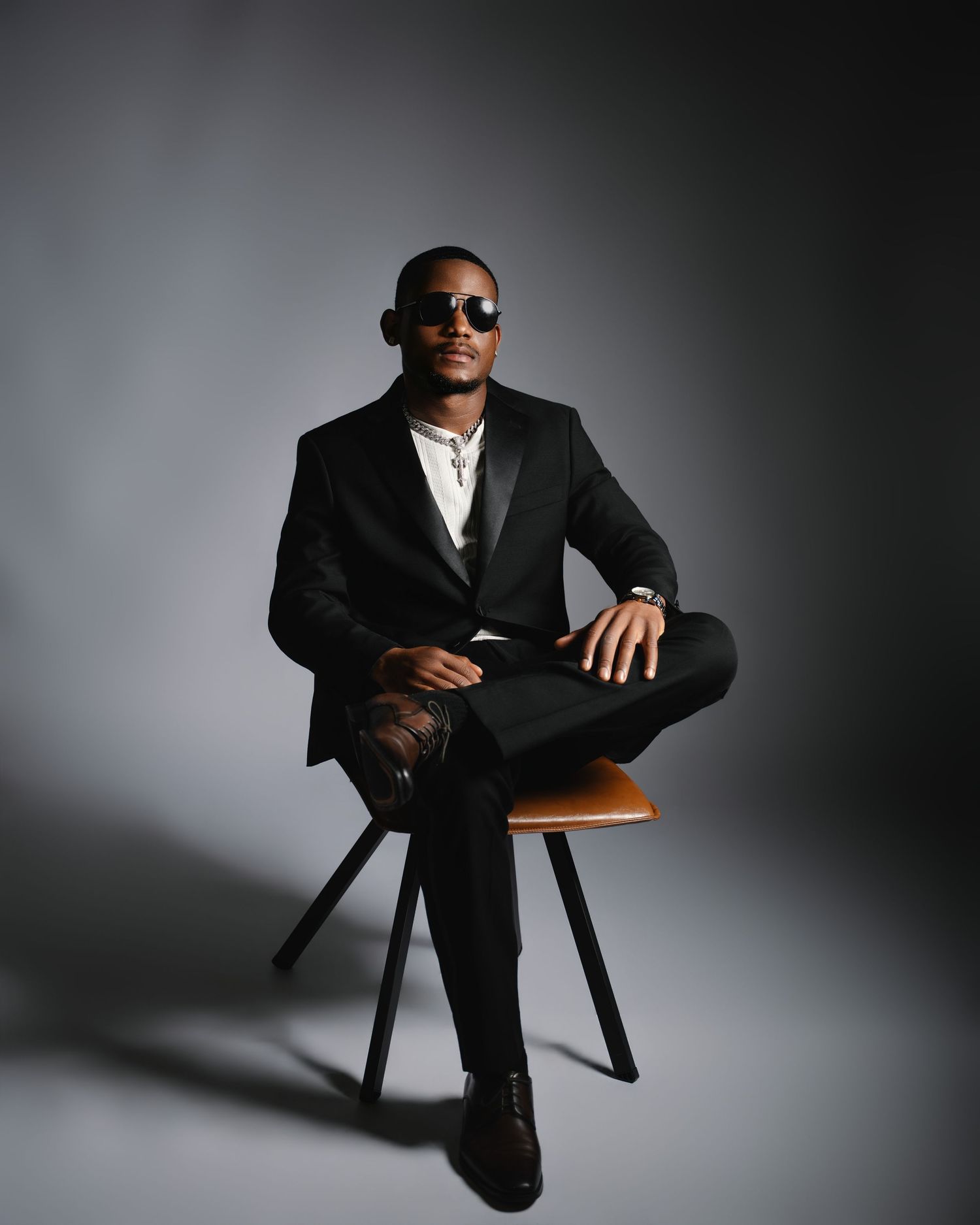 A well-dressed Black man sits cross-legged in a black tuxedo and sunglasses in a studio portrait.