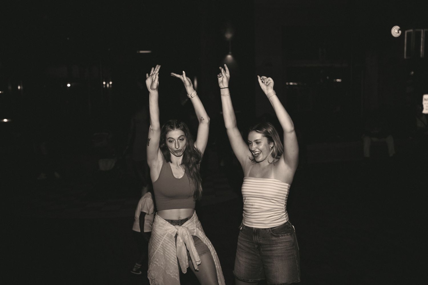 Black and white photo of two women celebrating with arms raised joyfully outdoors at night.