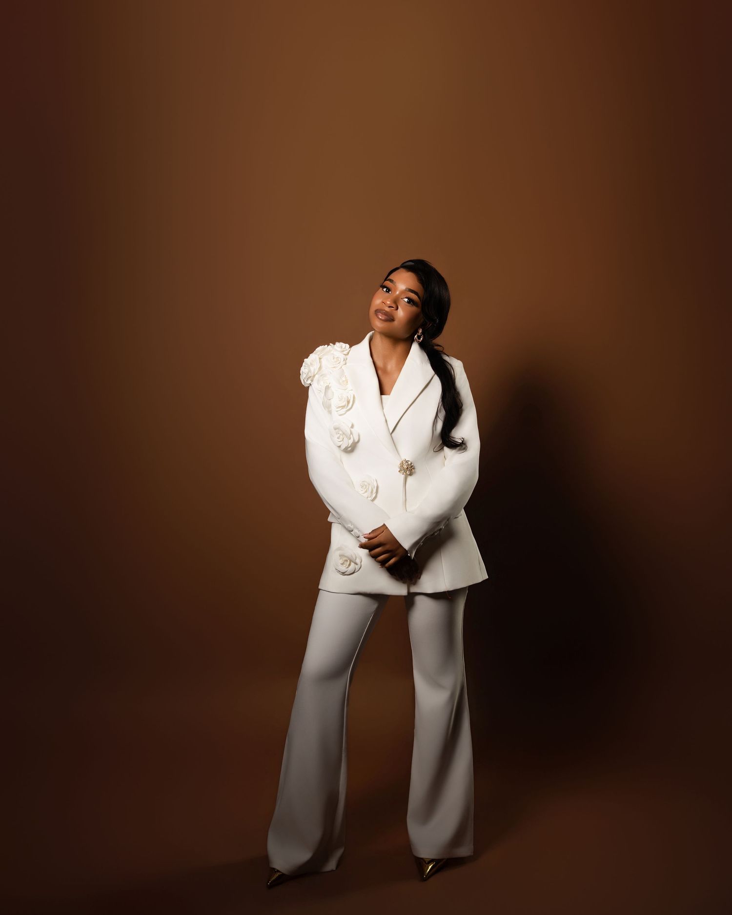 Young woman posing confidently in a white double-breasted blazer suit against a warm brown studio background.