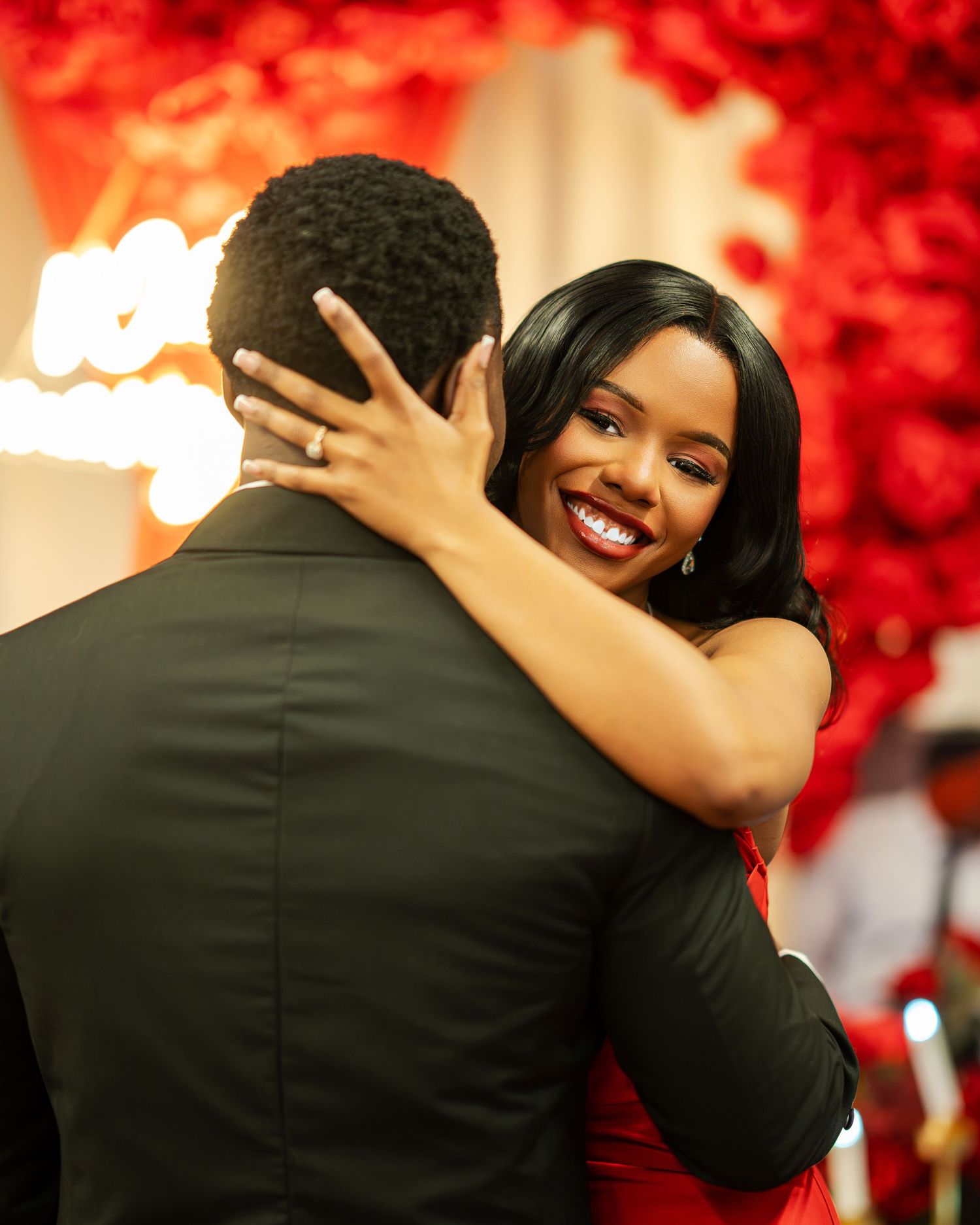 A joyful woman in a red dress hugs a man in a black suit surrounded by red roses at a romantic proposal.