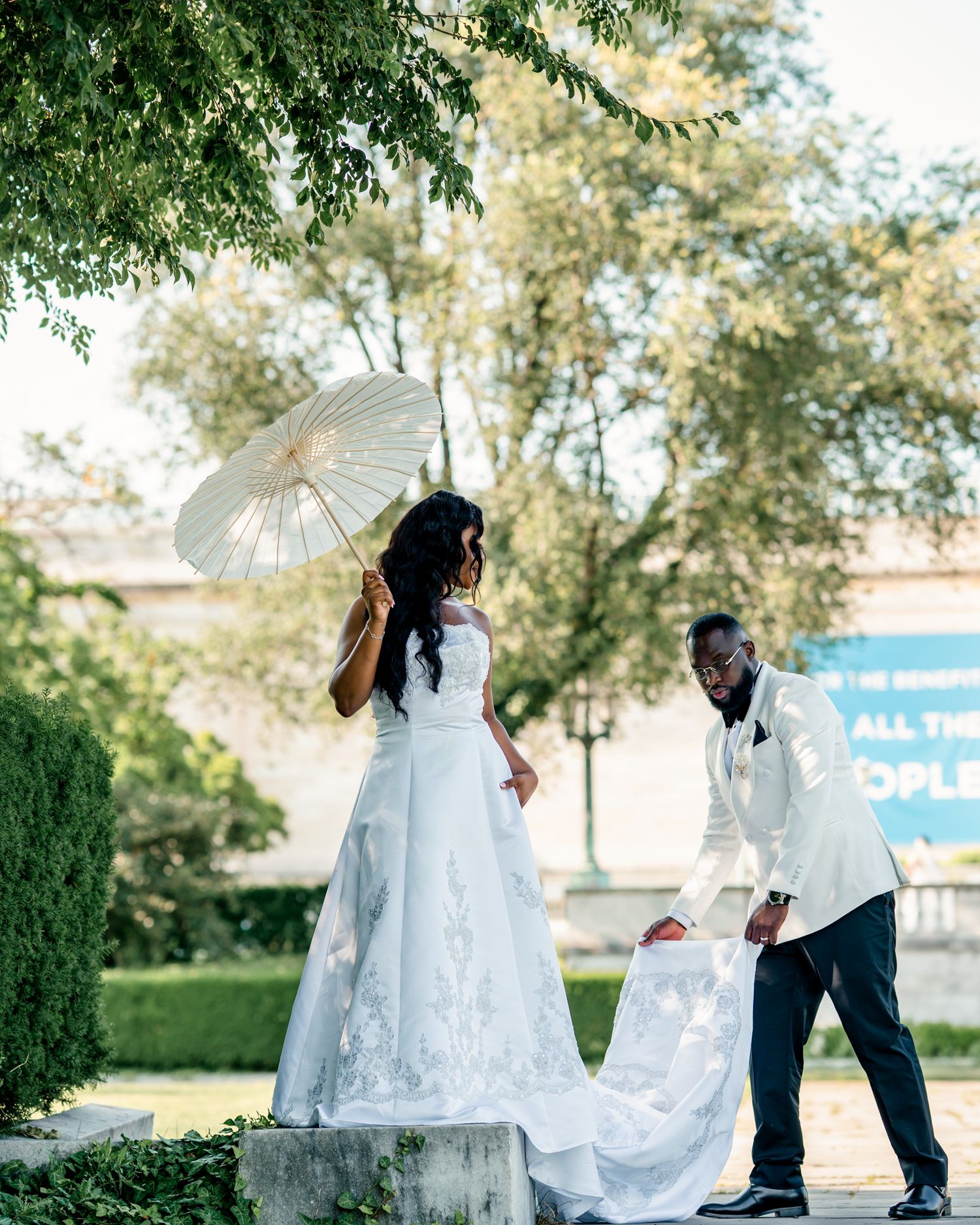 A groom in a white tuxedo gallantly helps arrange his bride's wedding gown train in a sunlit garden setting.