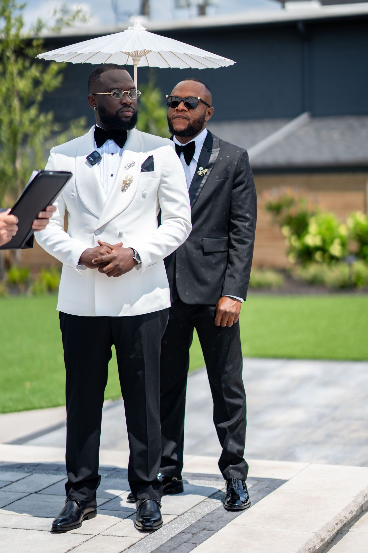 Two well-dressed groomsmen standing outdoors under a white parasol, one in white tuxedo jacket.