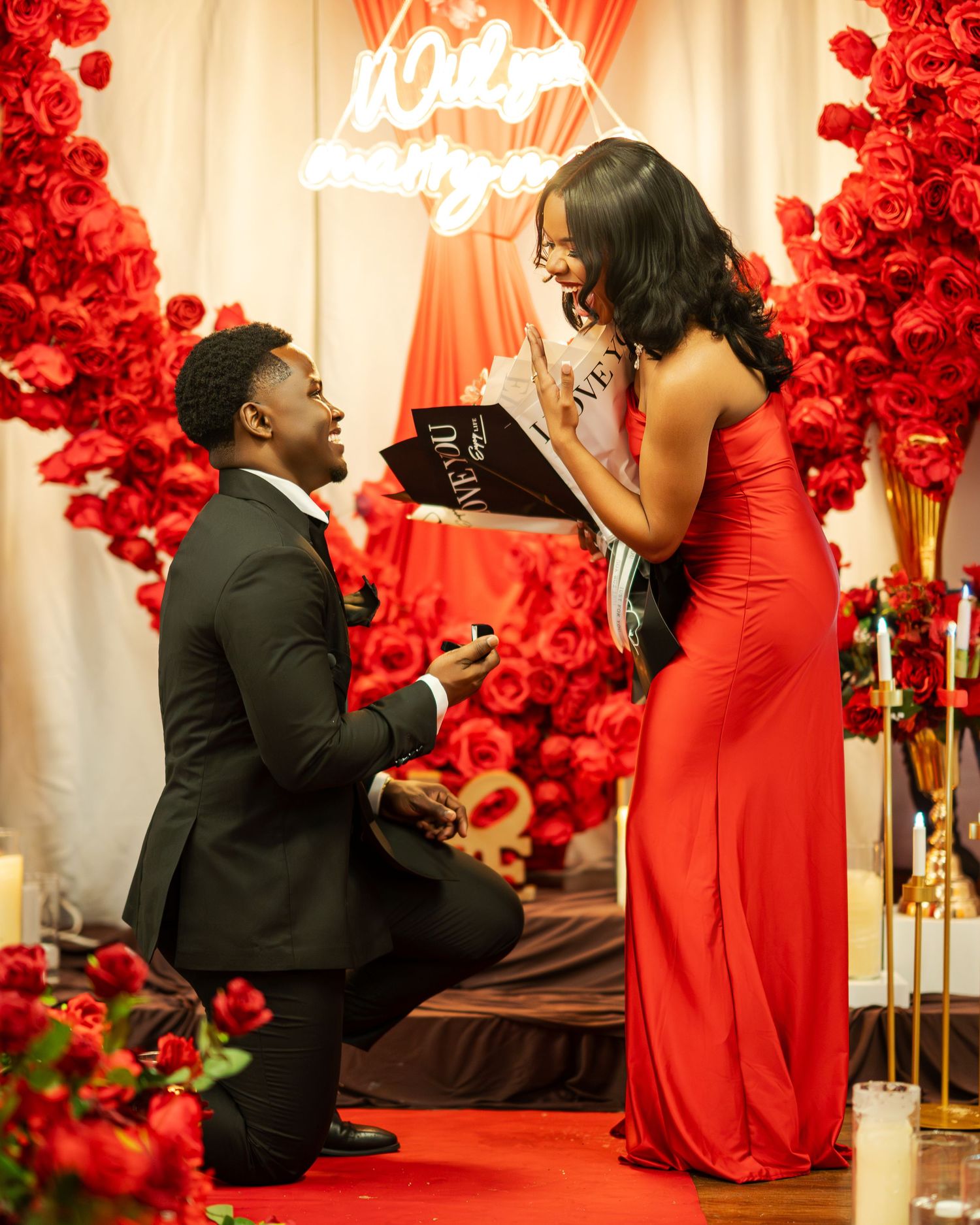 A man in a black suit proposes on one knee to a woman in a red gown surrounded by red roses and neon lights.