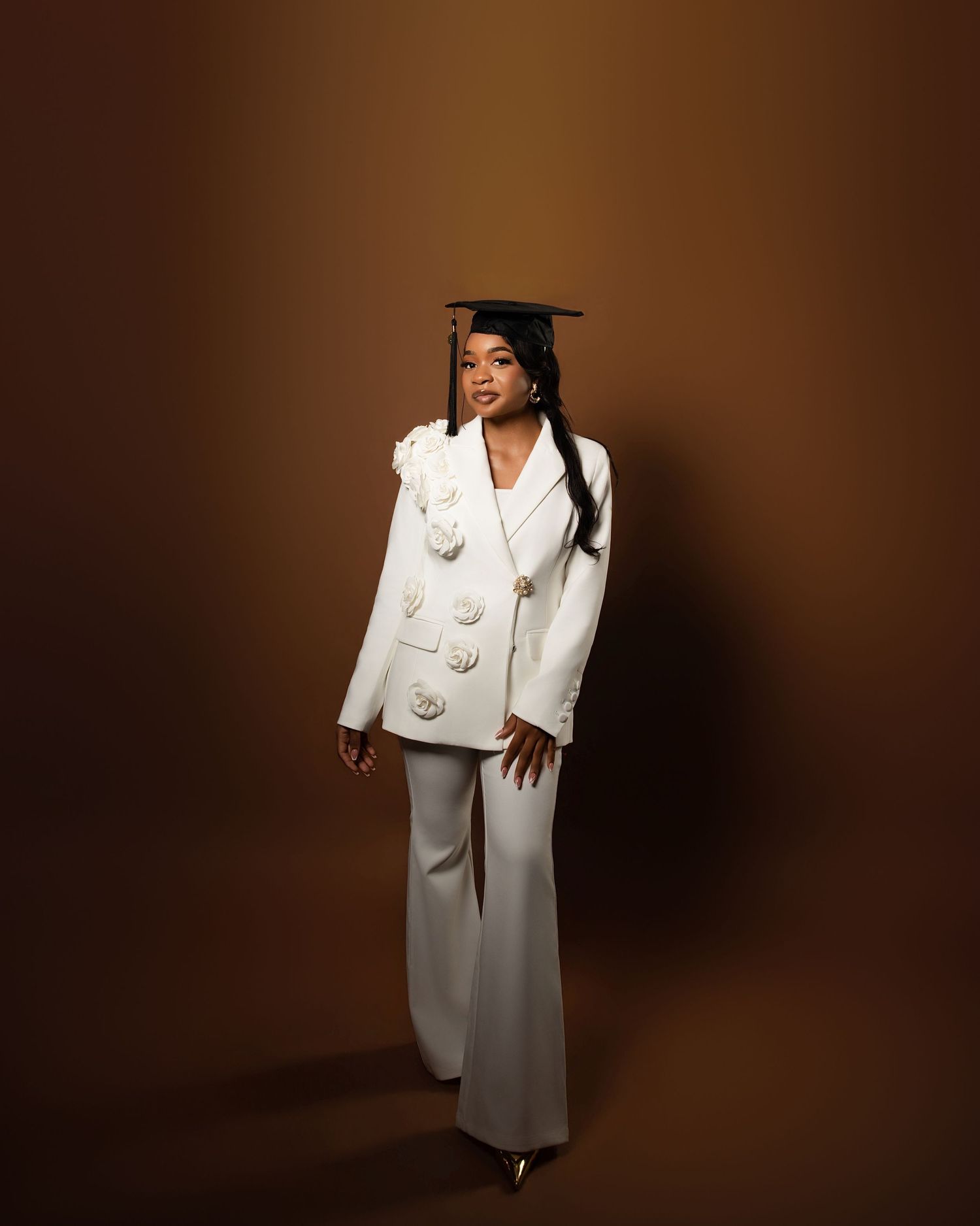 Graduate posing in white blazer suit and mortarboard cap against a warm brown studio background.