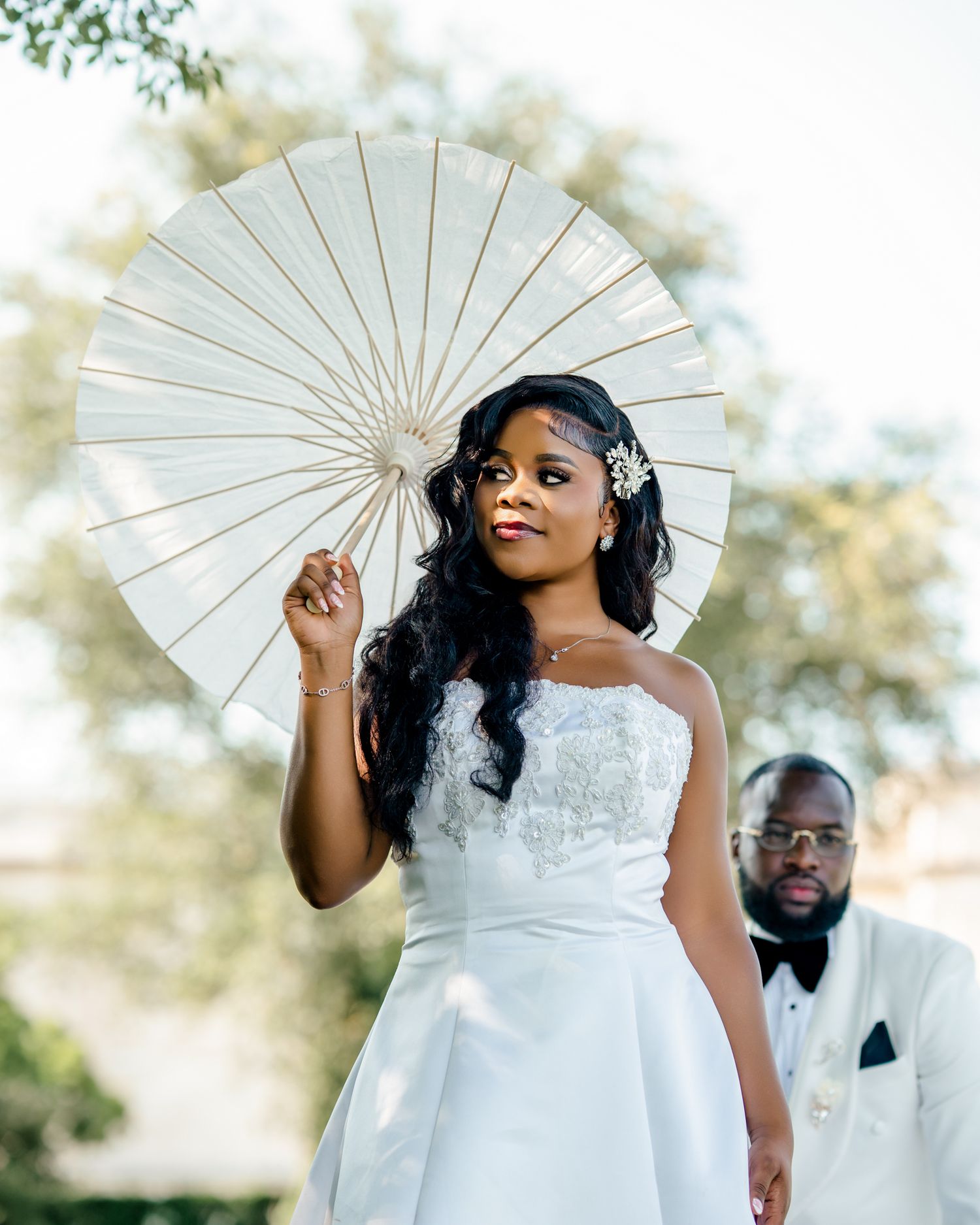 A radiant bride in a white strapless gown holds a parasol while her groom stands behind her outdoors.