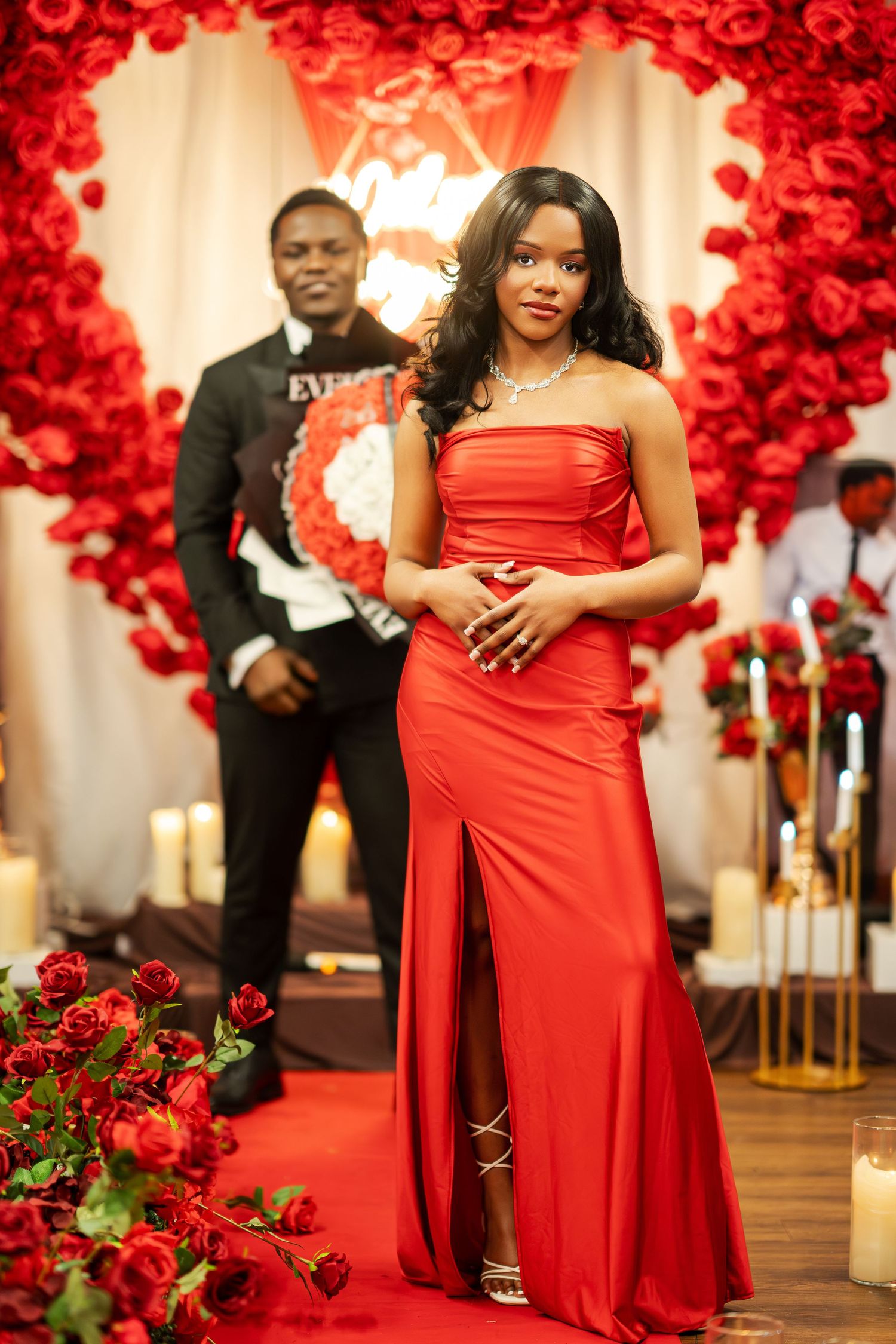 A woman in a red slit gown poses gracefully in front of a heart-shaped red rose wall with candles and a man behind her.
