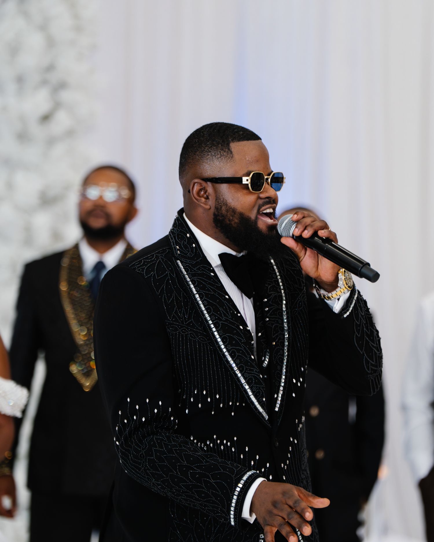 A man in a sparkling black tuxedo performs with a microphone at a wedding celebration.