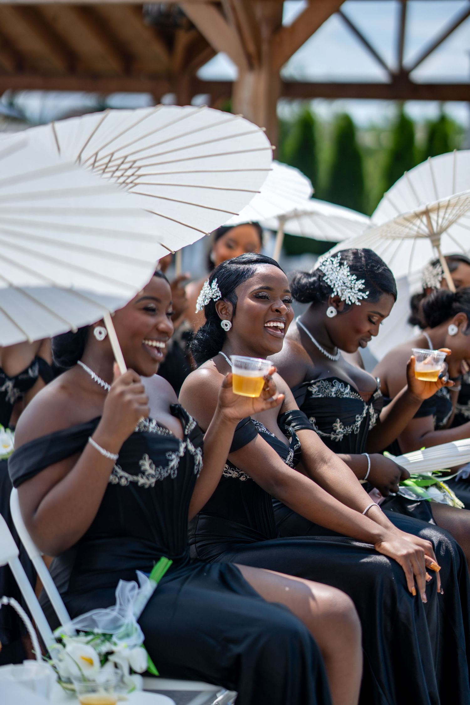 Bridesmaids in black dresses holding parasols and drinks while seated at outdoor wedding ceremony.