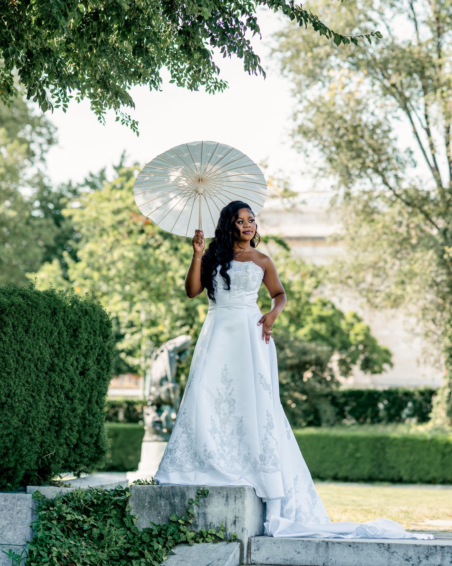 Bride in white strapless gown holding ivory parasol poses on stone steps in lush garden.