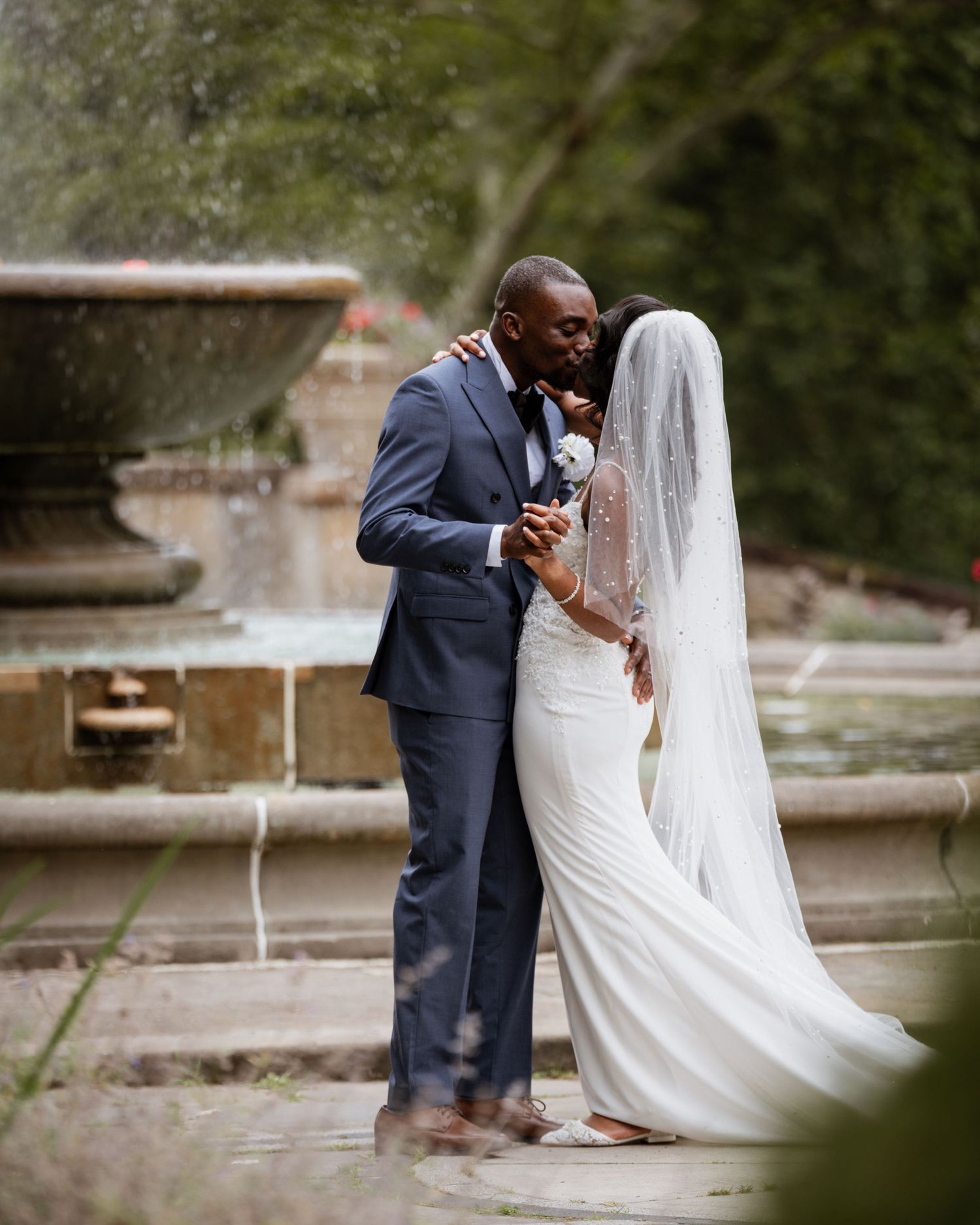 Bride and groom share romantic kiss near fountain, bride wearing white gown with long veil.