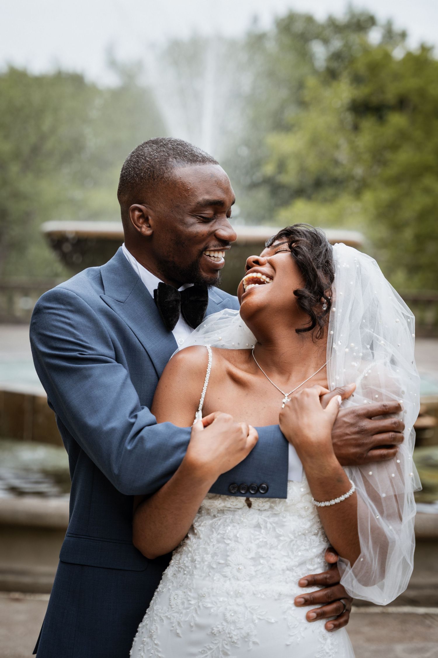 Joyful newlywed couple laughing and embracing near a fountain, groom in blue suit and bride in white wedding dress with veil.