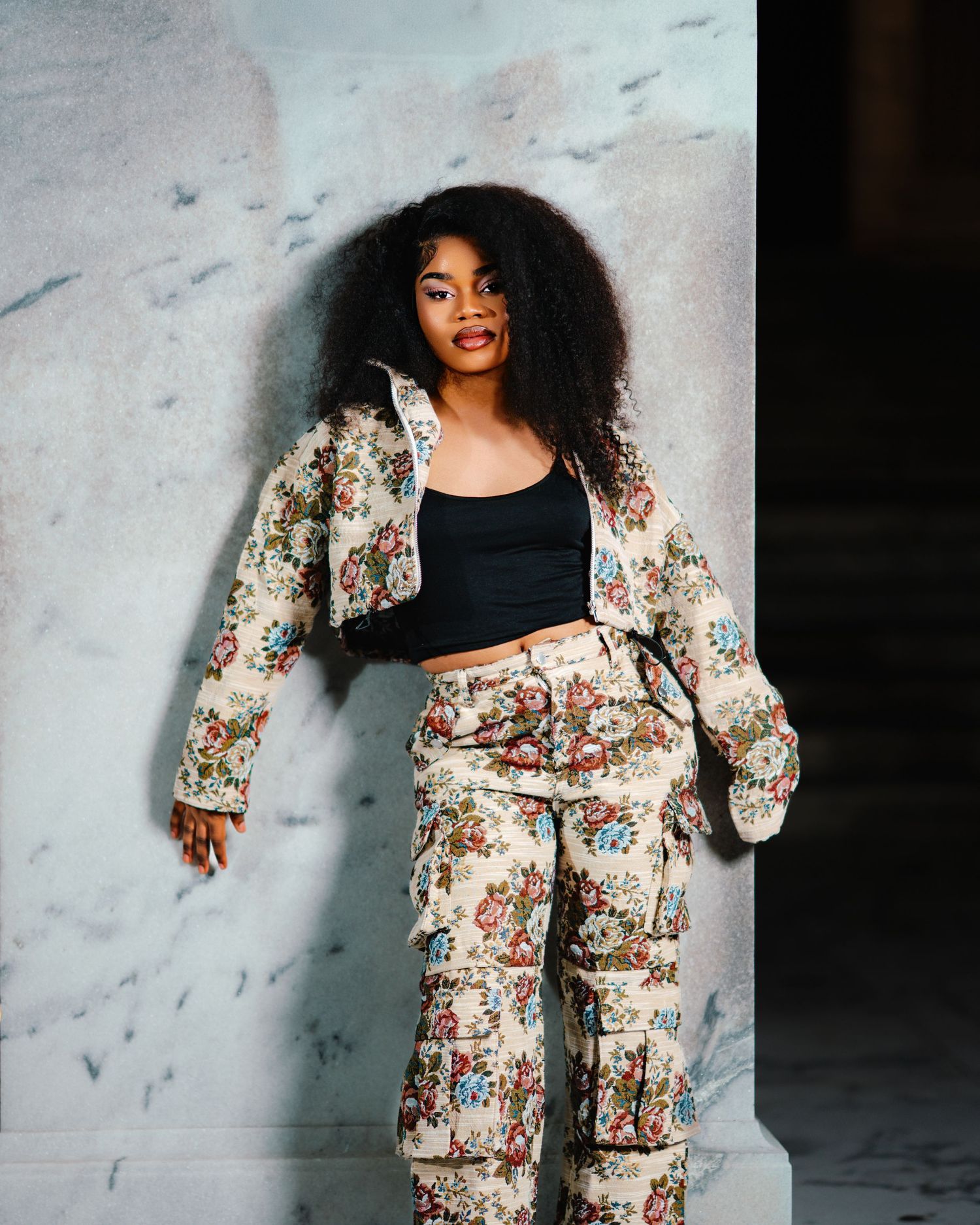 Young woman posing confidently in floral patterned jacket and matching cargo pants with black crop top against marble wall.
