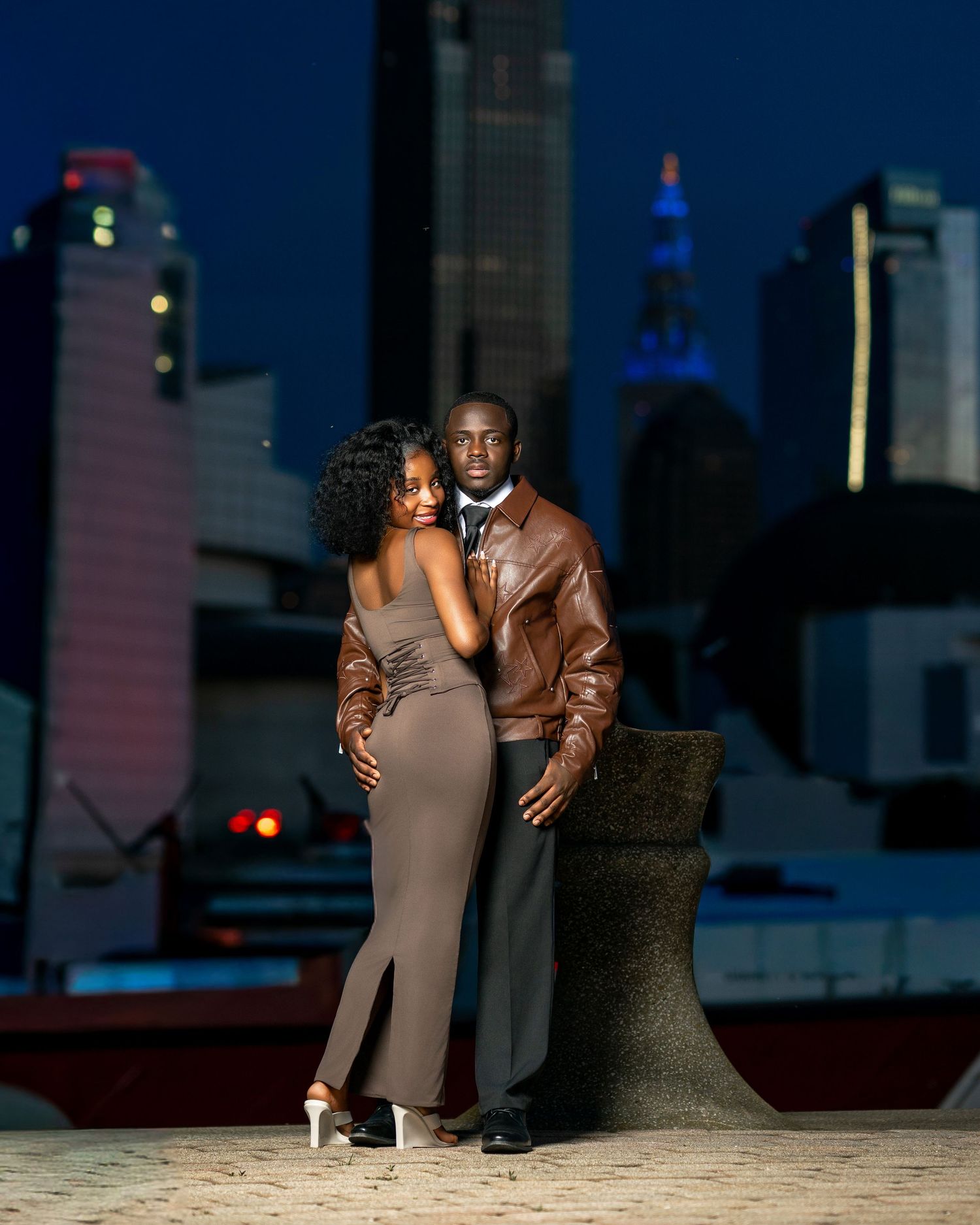Elegant couple posing at night with city skyline backdrop, woman in grey dress, man in brown leather jacket.