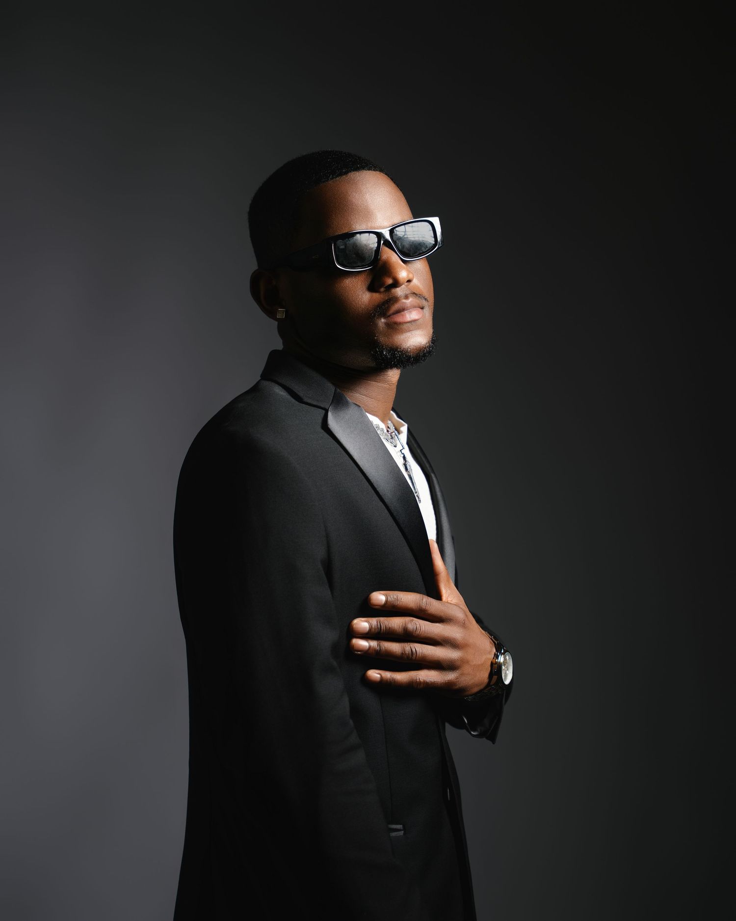 A confident Black man poses in a black suit and sunglasses against a dark gray studio background.