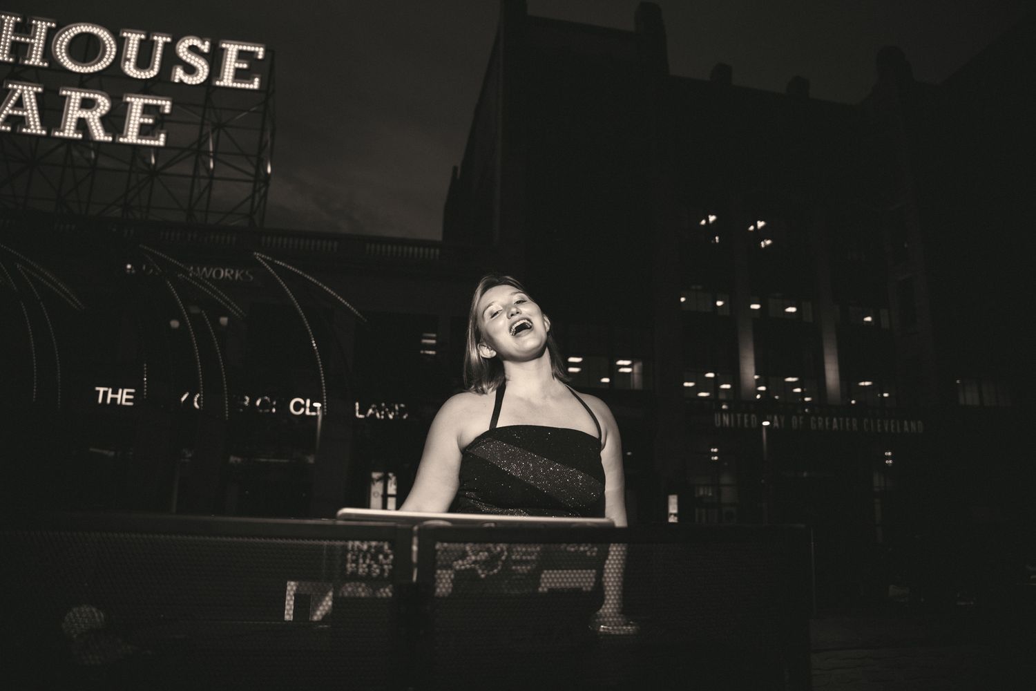Black and white photo of a woman laughing at an outdoor urban venue at night.