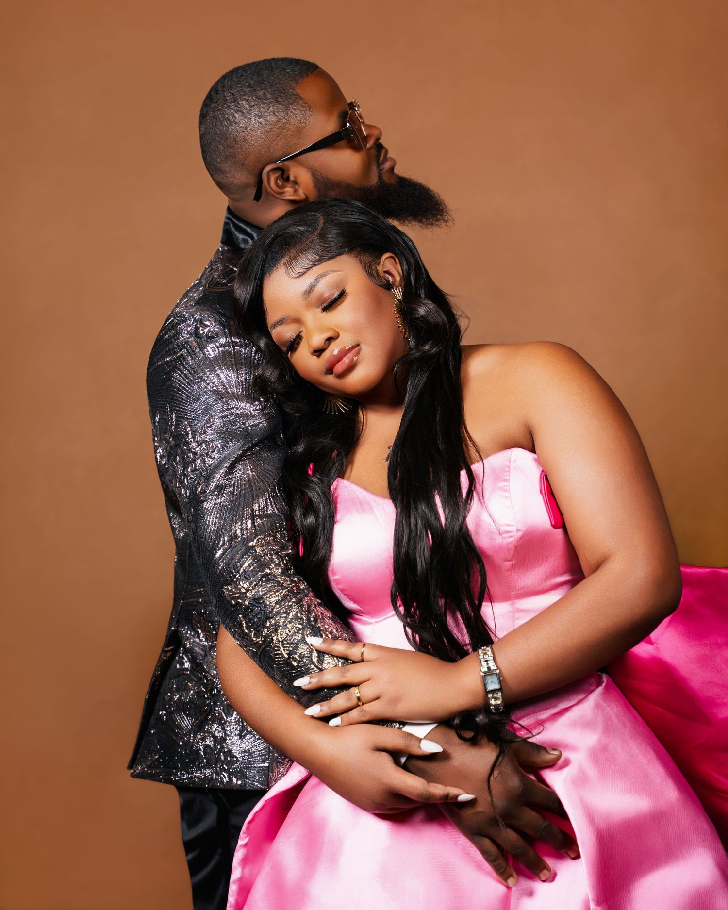 Romantic couple portrait with man embracing woman in pink satin strapless dress against warm brown background.