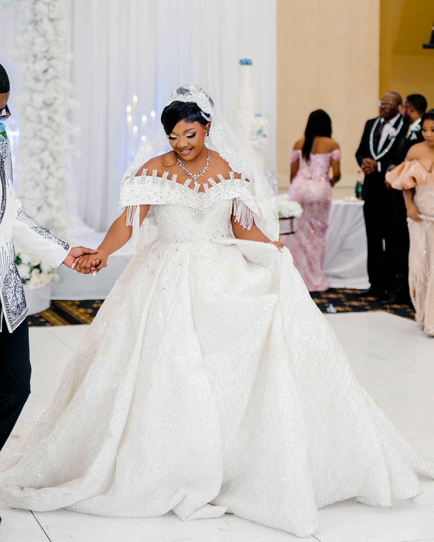 Bride in stunning off-shoulder ball gown with fringe details dances with groom at lavish wedding.