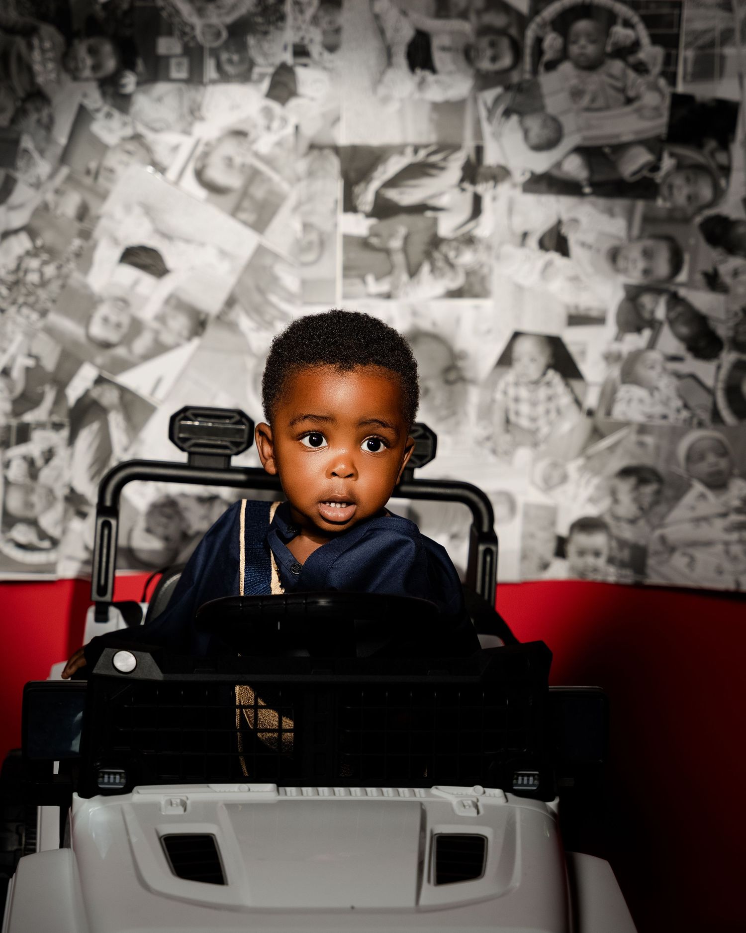 An adorable young boy in a navy hoodie sitting in a toy vehicle against a collage photo wall backdrop.